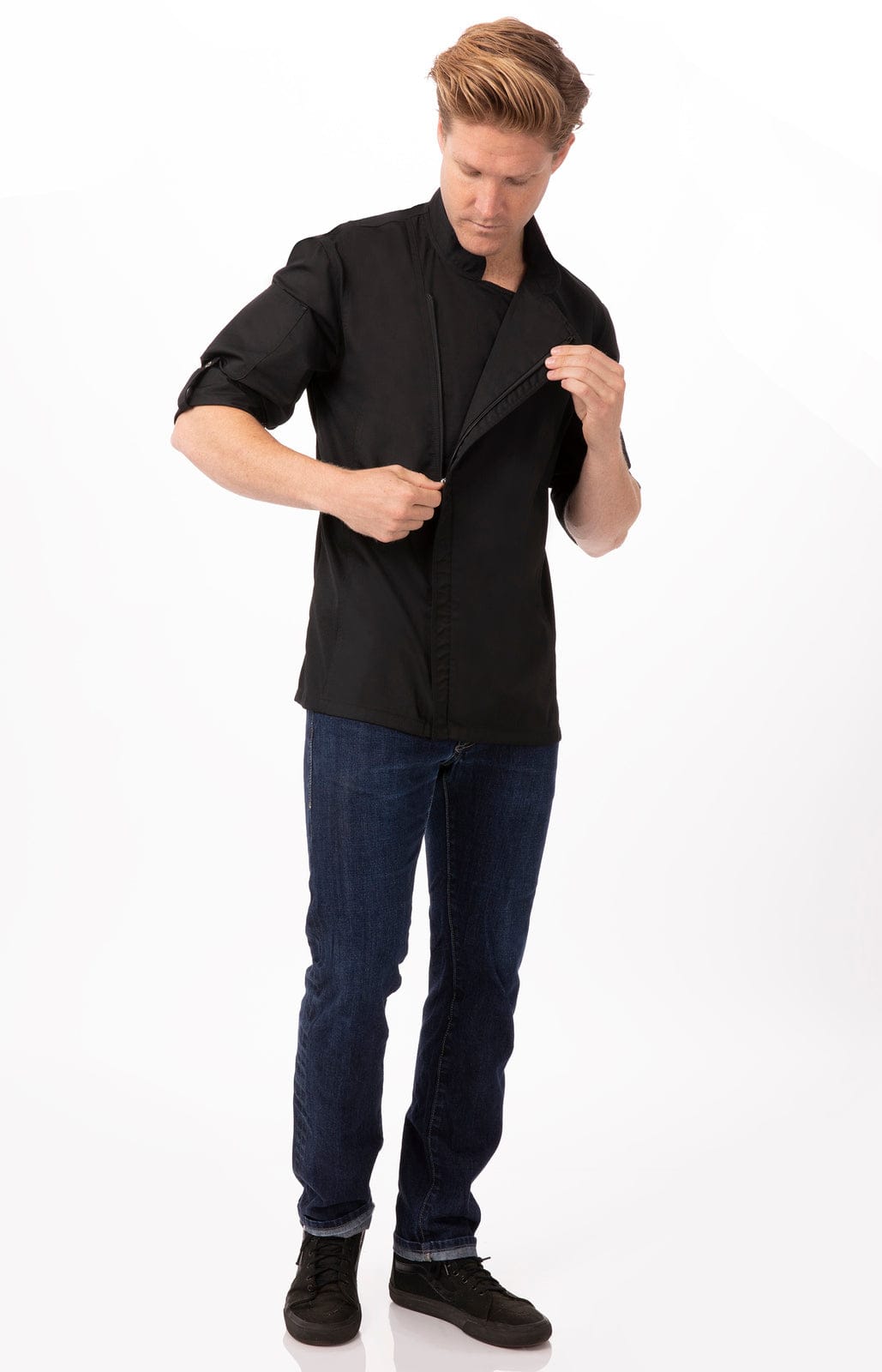 A man in a black chef jacket adjusts the front, paired with blue jeans and black shoes. The jacket has a logo.