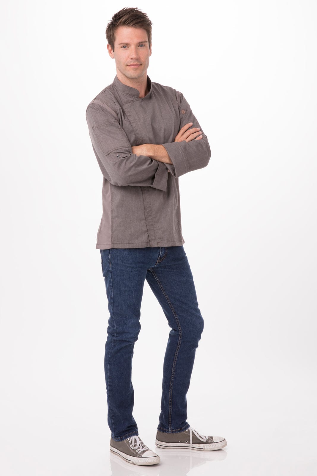 A man wearing a grey chef jacket with long sleeves, paired with blue jeans and casual shoes, stands against a light background.