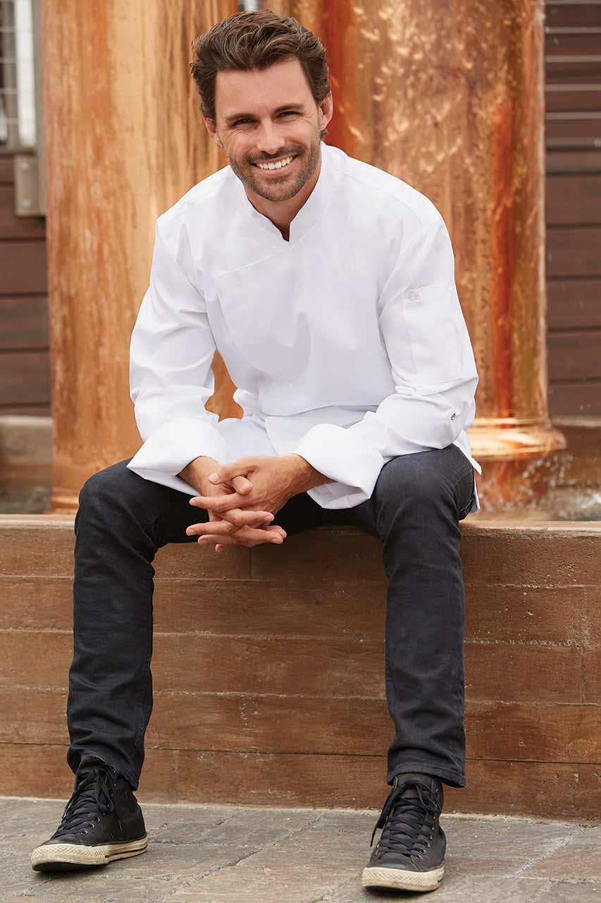A man wearing a white chef jacket and black pants sits casually, smiling, against a rustic background.