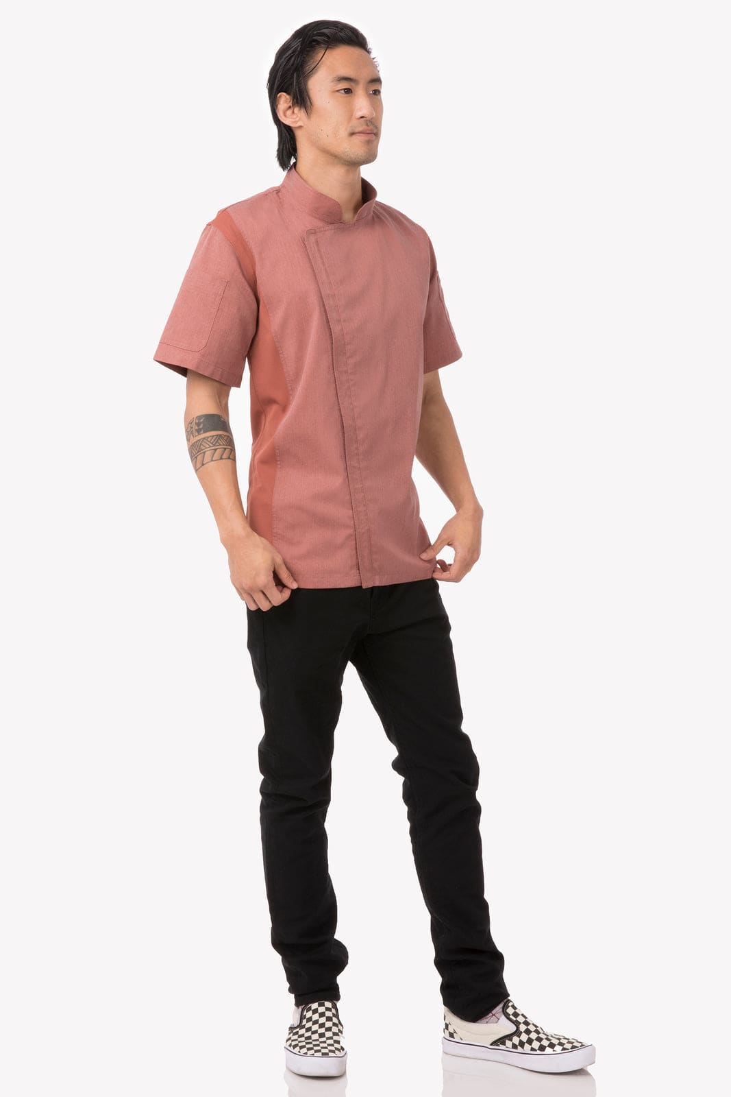 A man stands wearing a short-sleeved, rose-coloured chef jacket paired with black pants and checkered shoes.