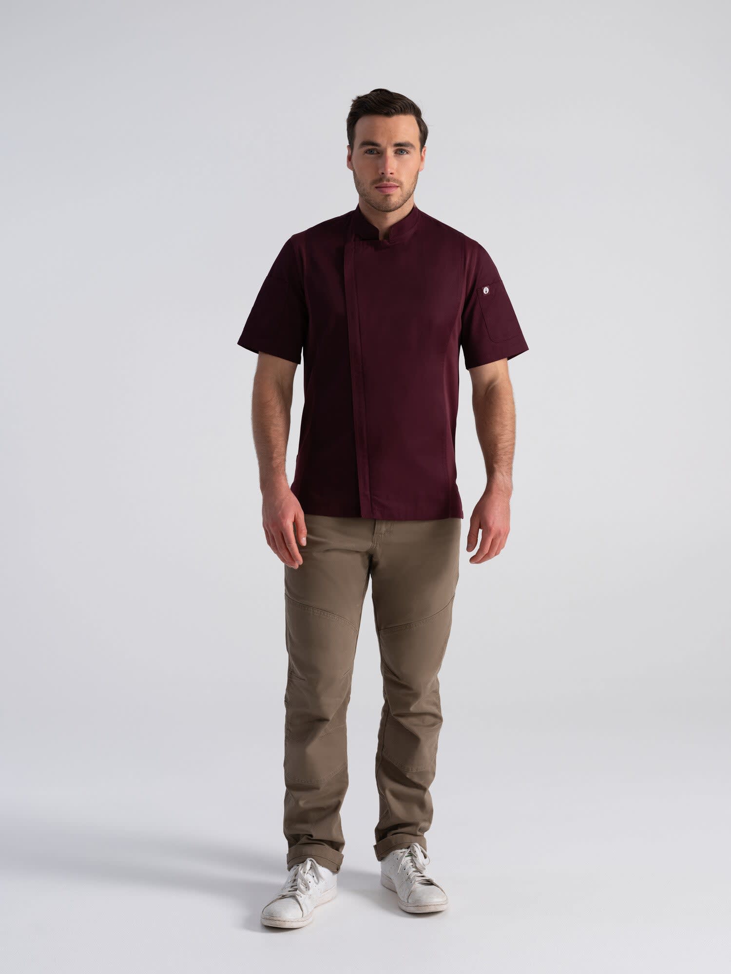 A man stands wearing a burgundy chef jacket with short sleeves, paired with beige pants and white shoes. The jacket has a logo.