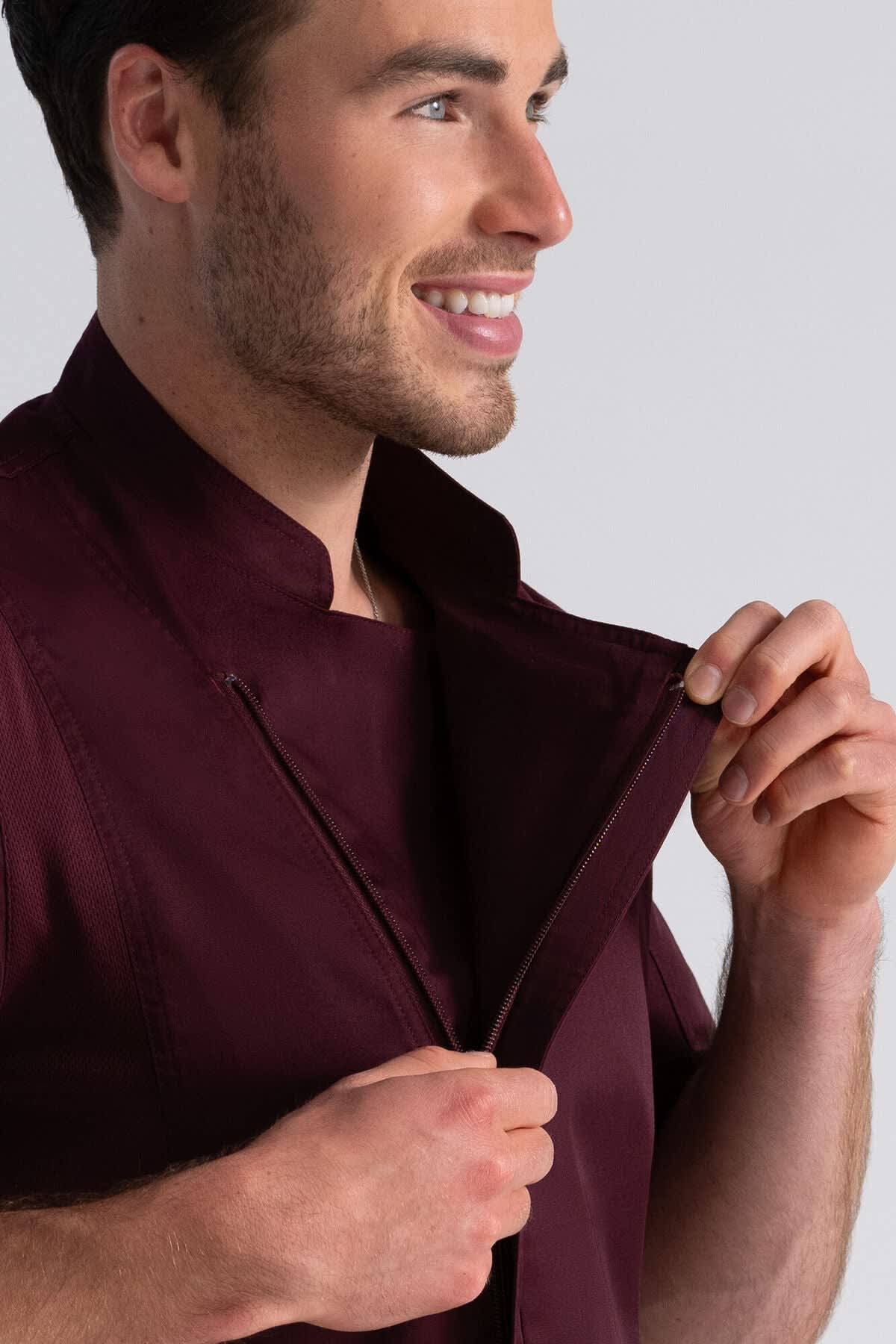 A man in a burgundy chef jacket is smiling while adjusting the collar. The jacket has a logo on it.