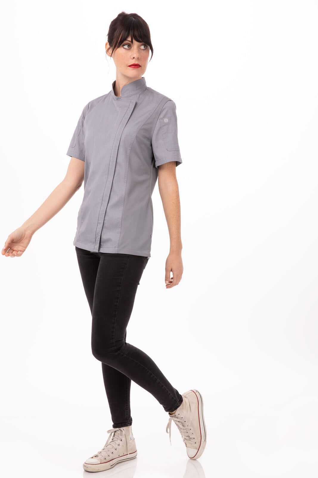 A woman in a grey chef jacket paired with black pants and sneakers, standing in a relaxed pose. The jacket features a logo.