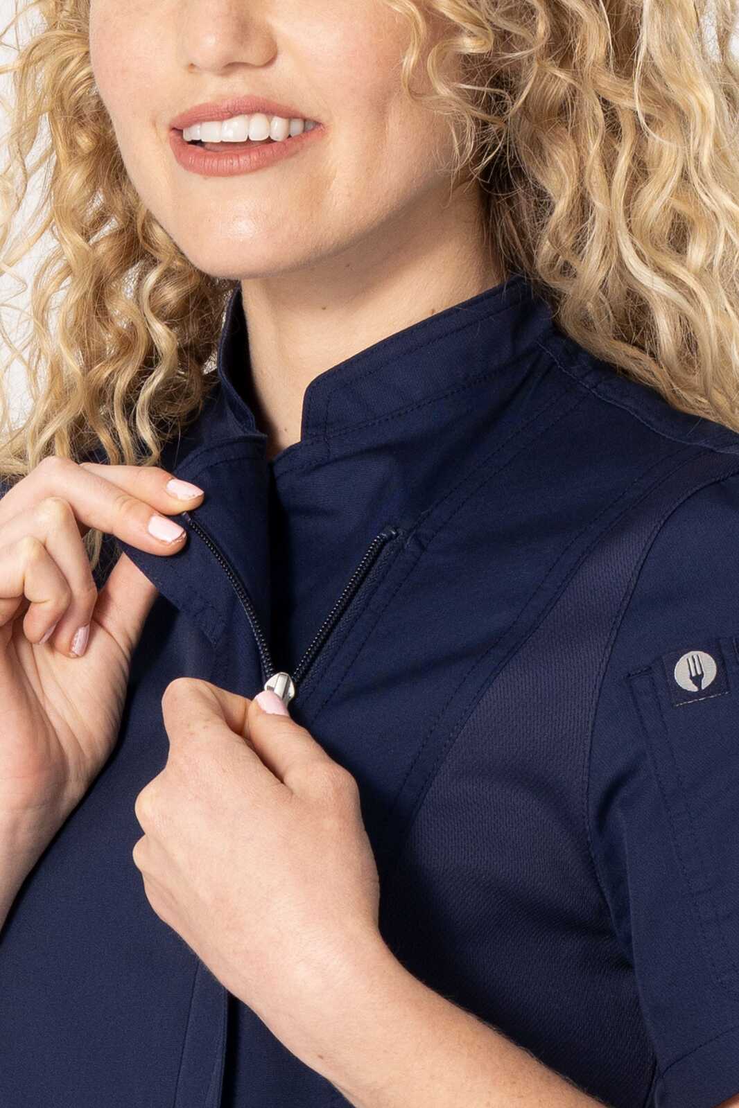 A close-up of a woman in a navy chef jacket, smiling and adjusting the zip collar. The jacket features a logo.
