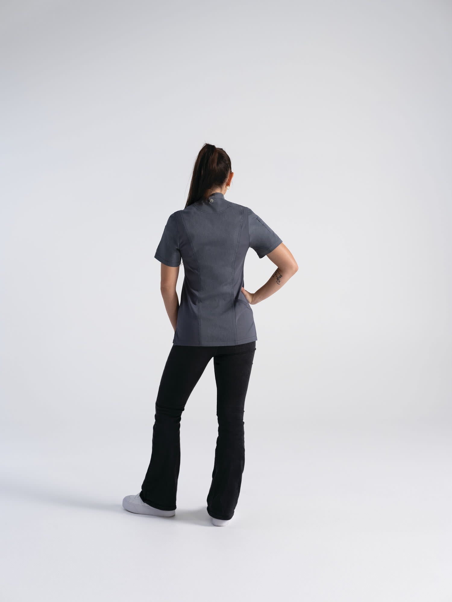 A women's chef jacket in grey, worn by a model facing away, paired with black pants. The jacket has a subtle logo.