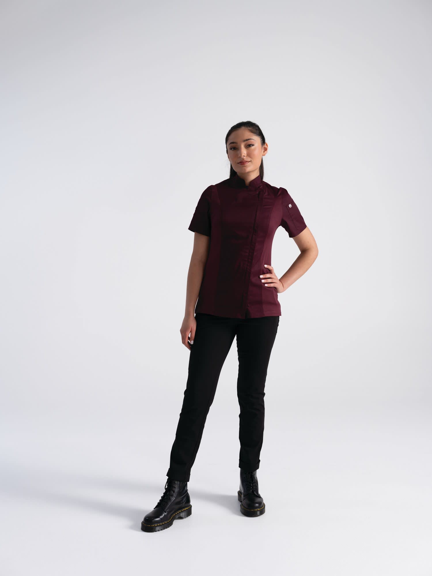 A woman stands in a burgundy women's chef jacket paired with black pants and boots, featuring a logo on the sleeve.