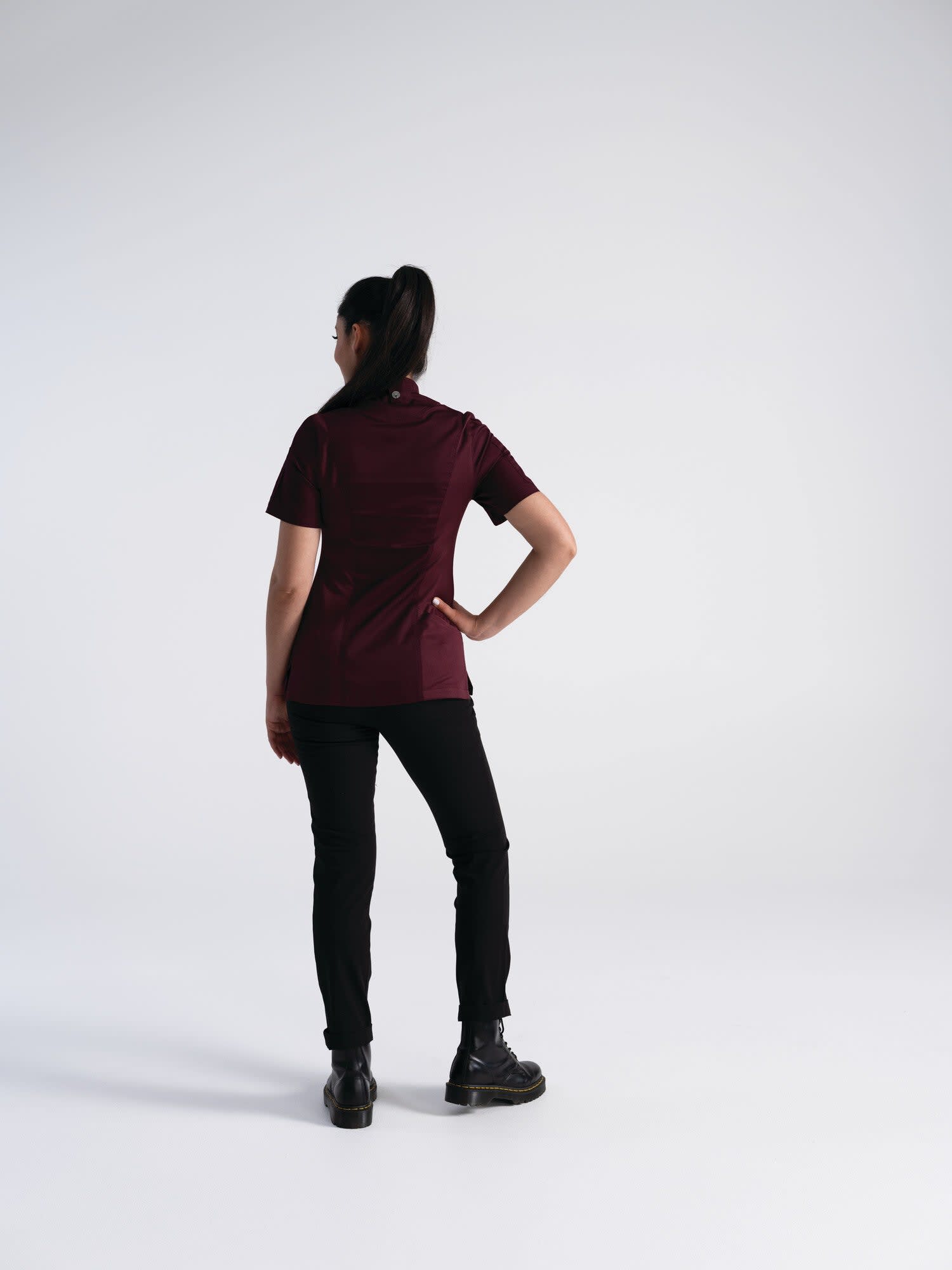 A woman wearing a burgundy chef jacket and black pants stands with her back to the camera in a studio setting.