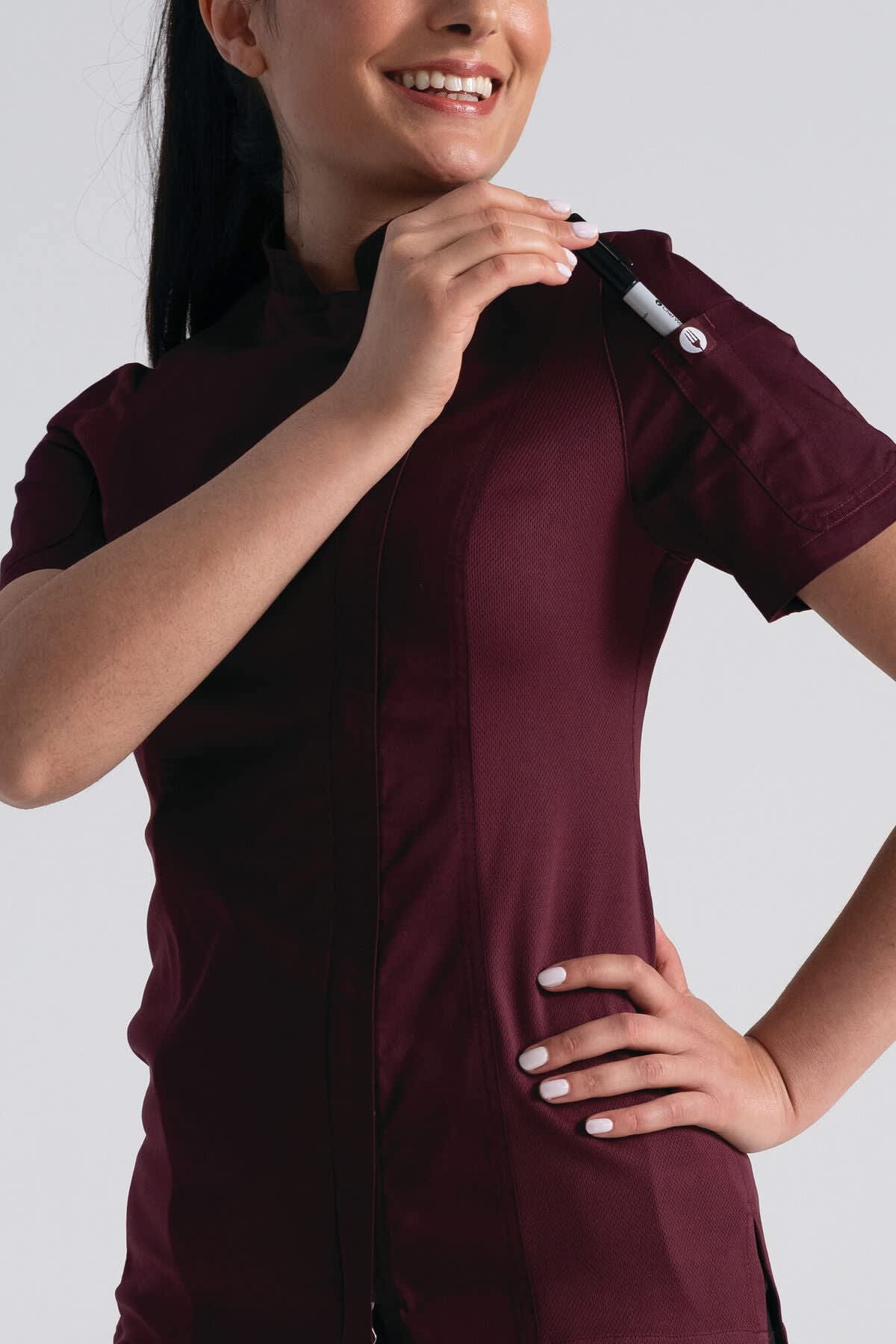 A woman models a burgundy chef jacket, featuring short sleeves and a side pocket for a pen. The garment has a logo.