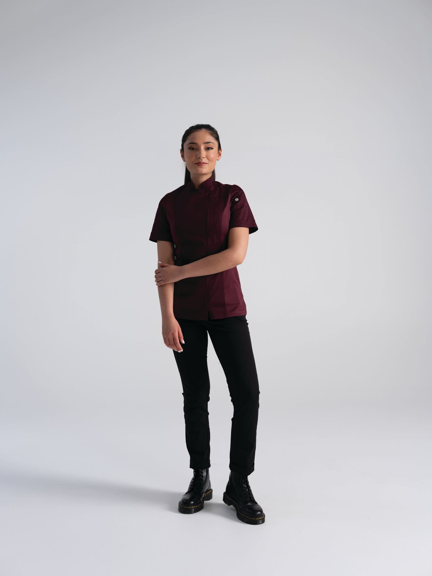 A women's chef jacket in burgundy displayed on a model, paired with black trousers and boots. The jacket features a logo.