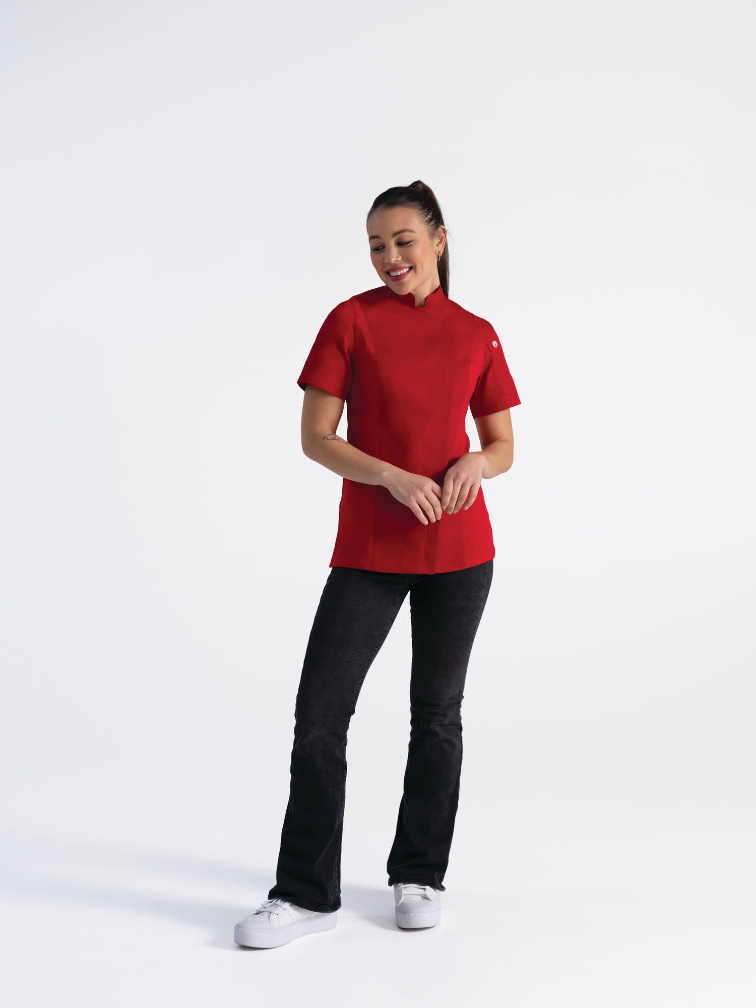 A woman wearing a red chef jacket and black pants, standing confidently against a white background. The jacket has a logo.