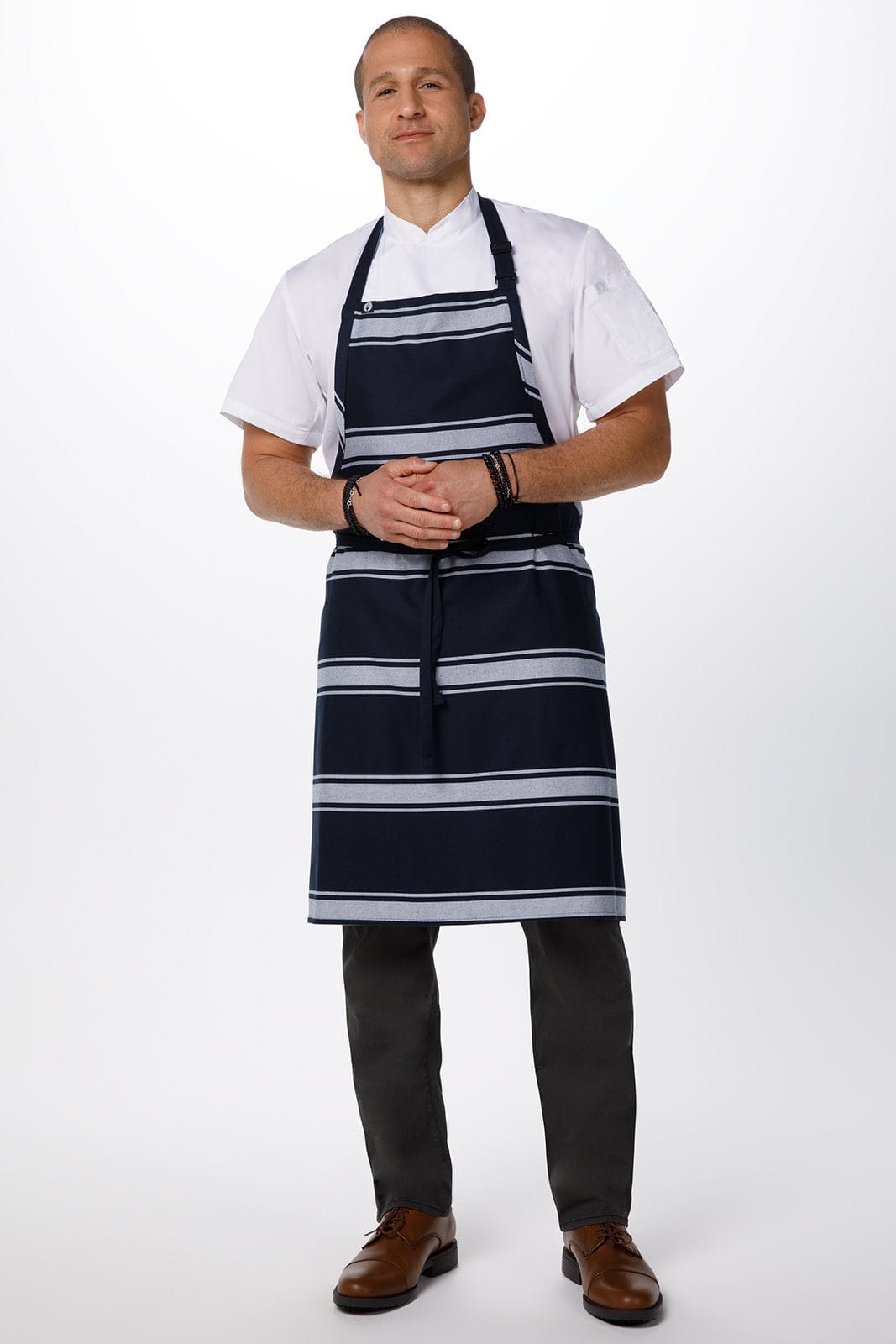 A man wearing a navy butcher stripe bib apron over a white shirt, paired with dark trousers and brown shoes.