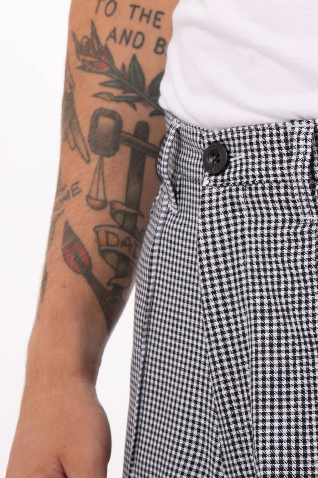 Black and white check patterned chef pants with a button closure and visible side pocket. Features a tattooed arm in the background.