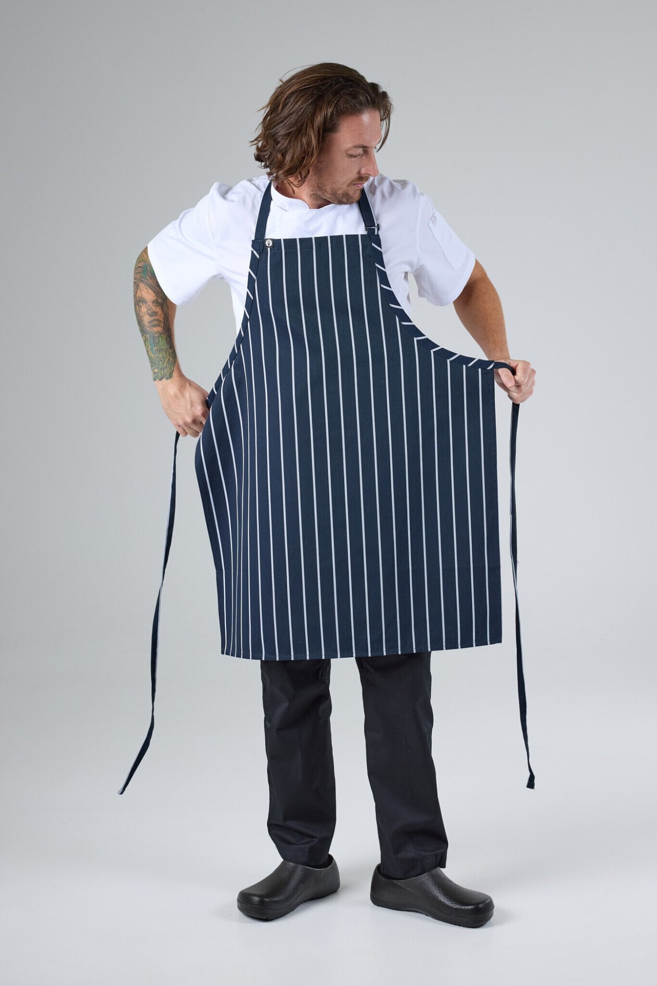 A navy and white striped bib apron is being worn by a person, featuring ties on the side. The person is dressed in a white top and black trousers.