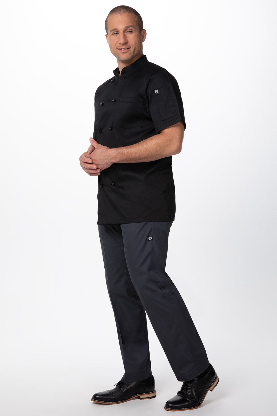 A man wears a black chef jacket with short sleeves, paired with grey pants and black shoes, standing confidently.
