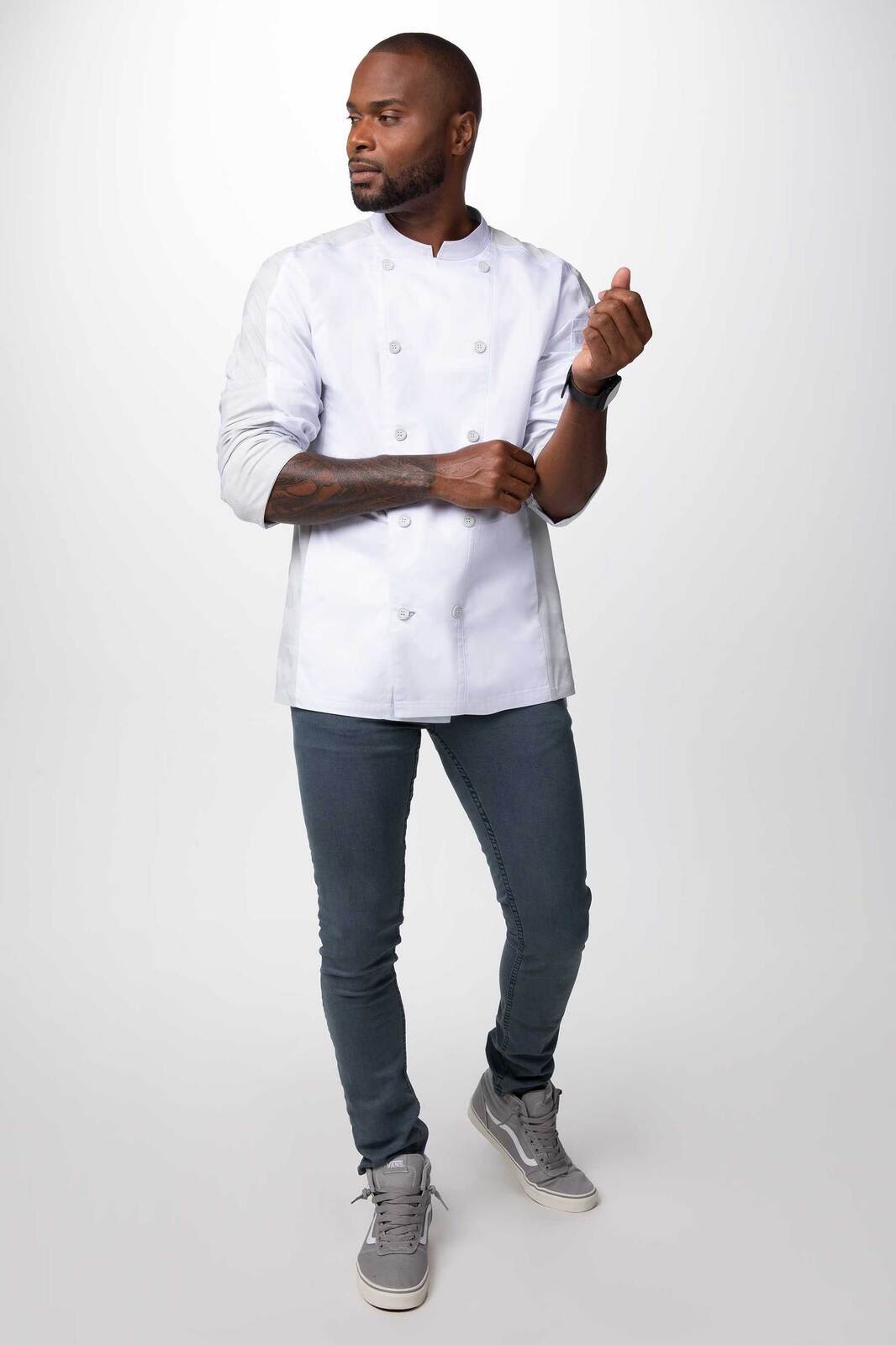A man wearing a white chef jacket and grey jeans, standing with a relaxed posture against a light background.