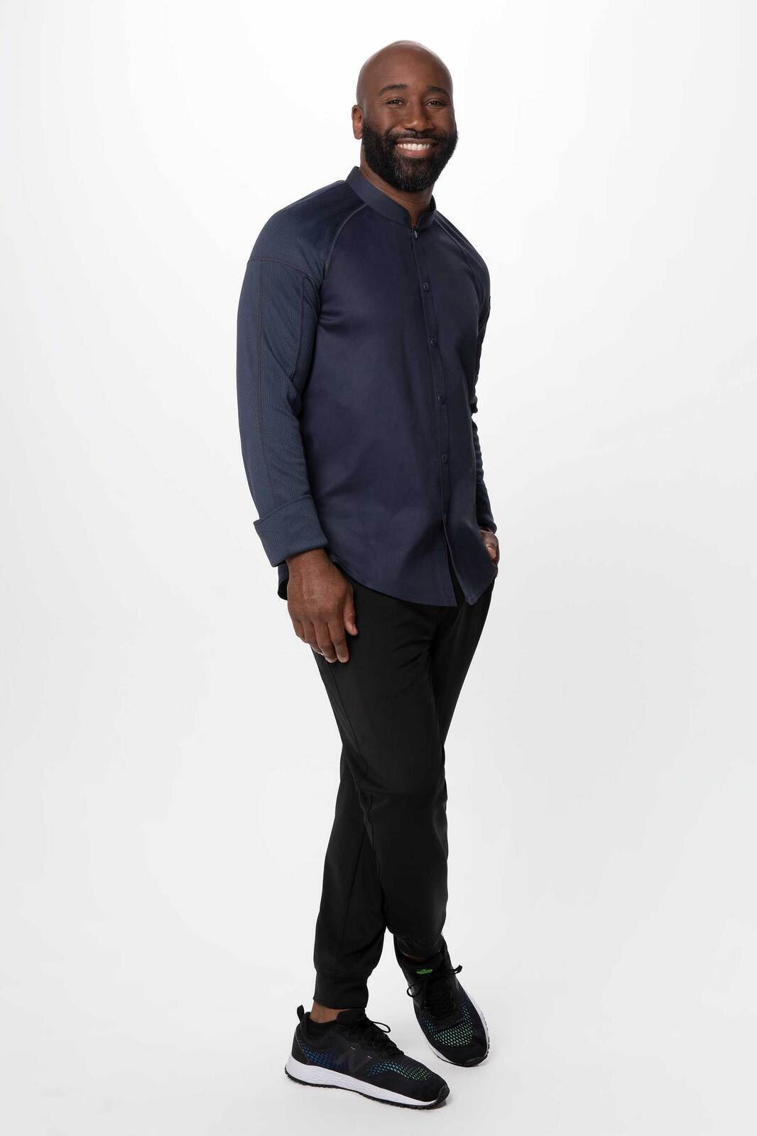 A man wearing a navy Tulum Men's Chef Jacket paired with black pants and sneakers, smiling in a neutral background.