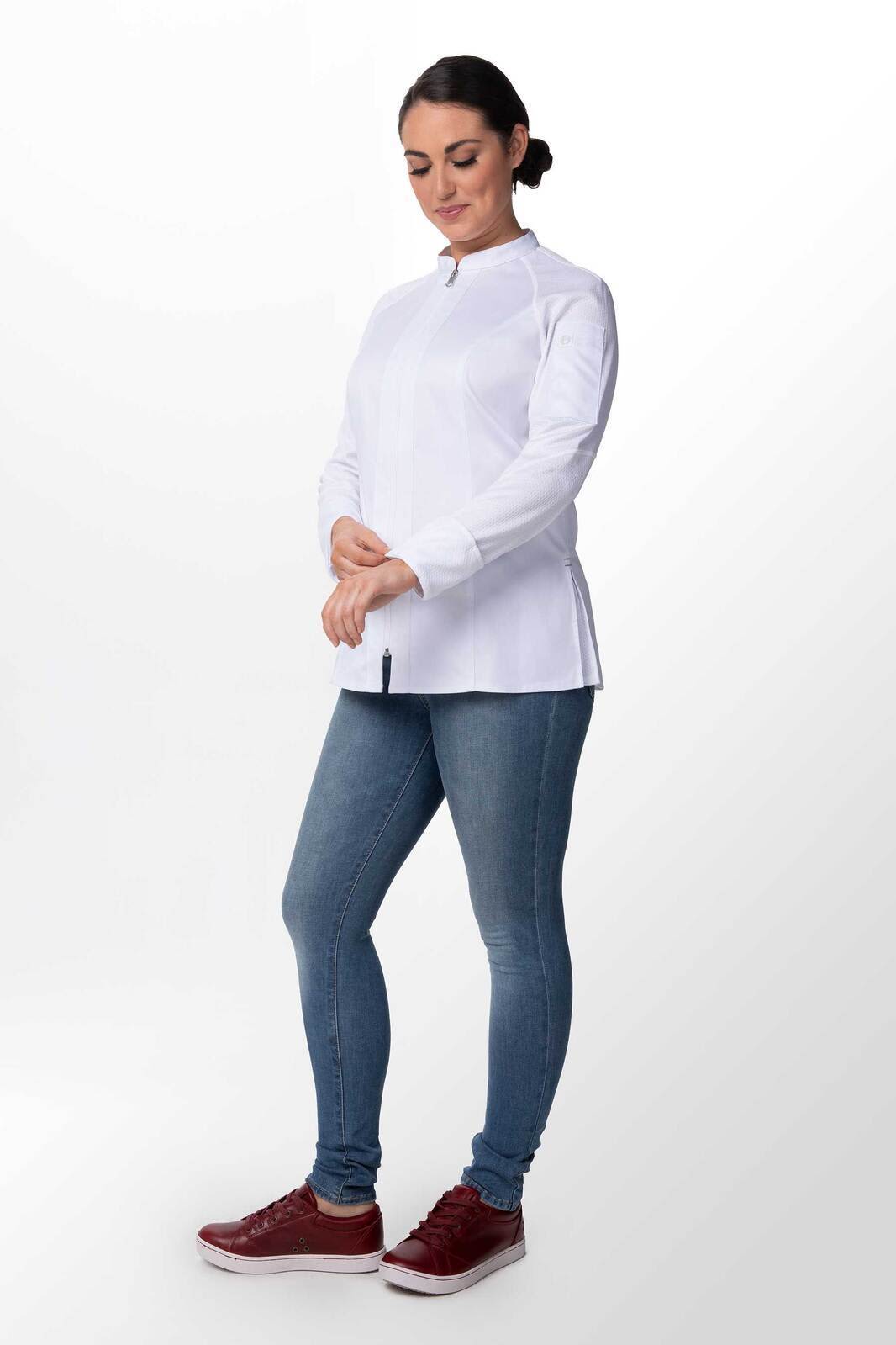 A woman is wearing a white chef jacket paired with blue jeans and red shoes, standing with one hand on her wrist.