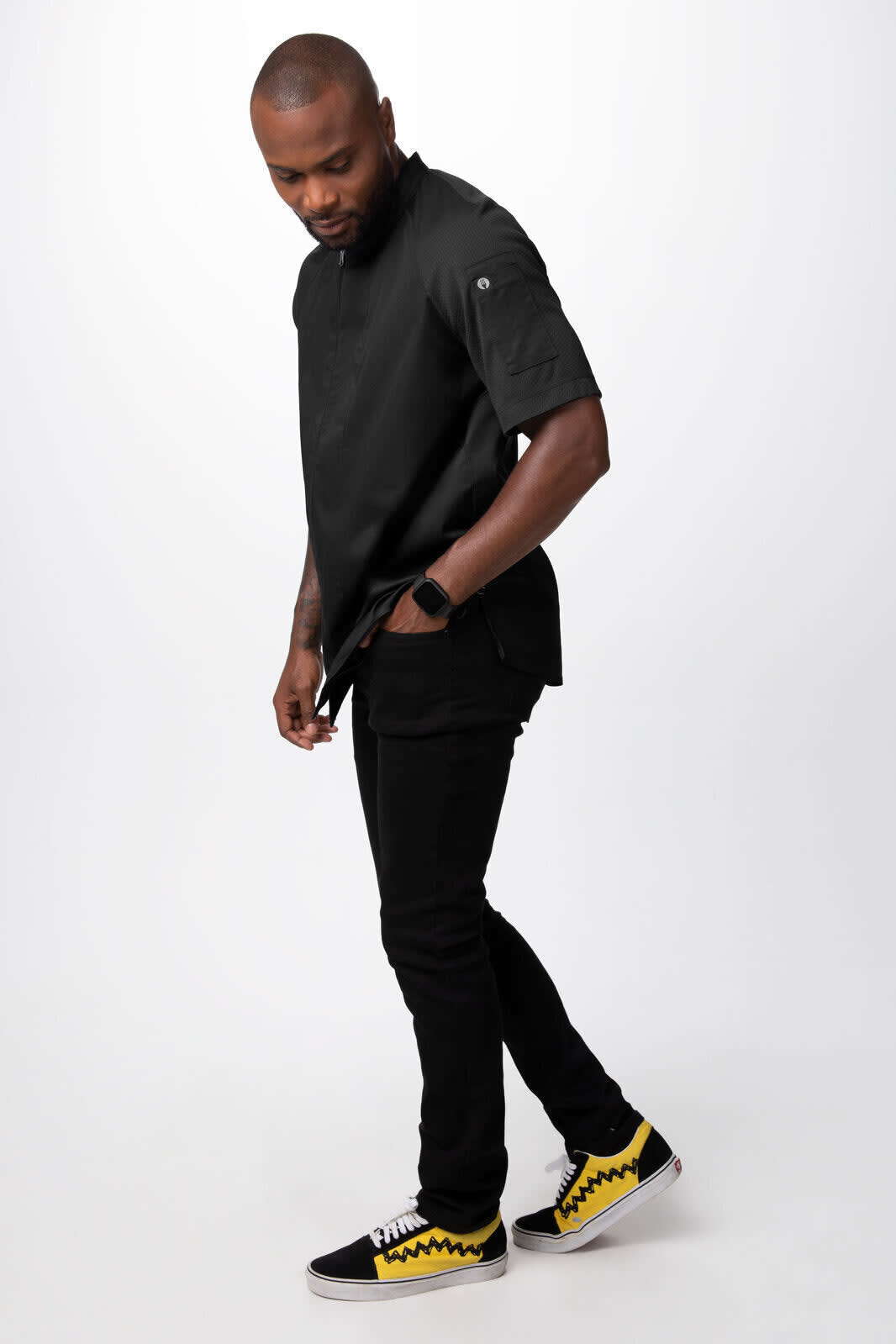 A black men's chef jacket is worn by a man, paired with black pants and yellow-accented sneakers.