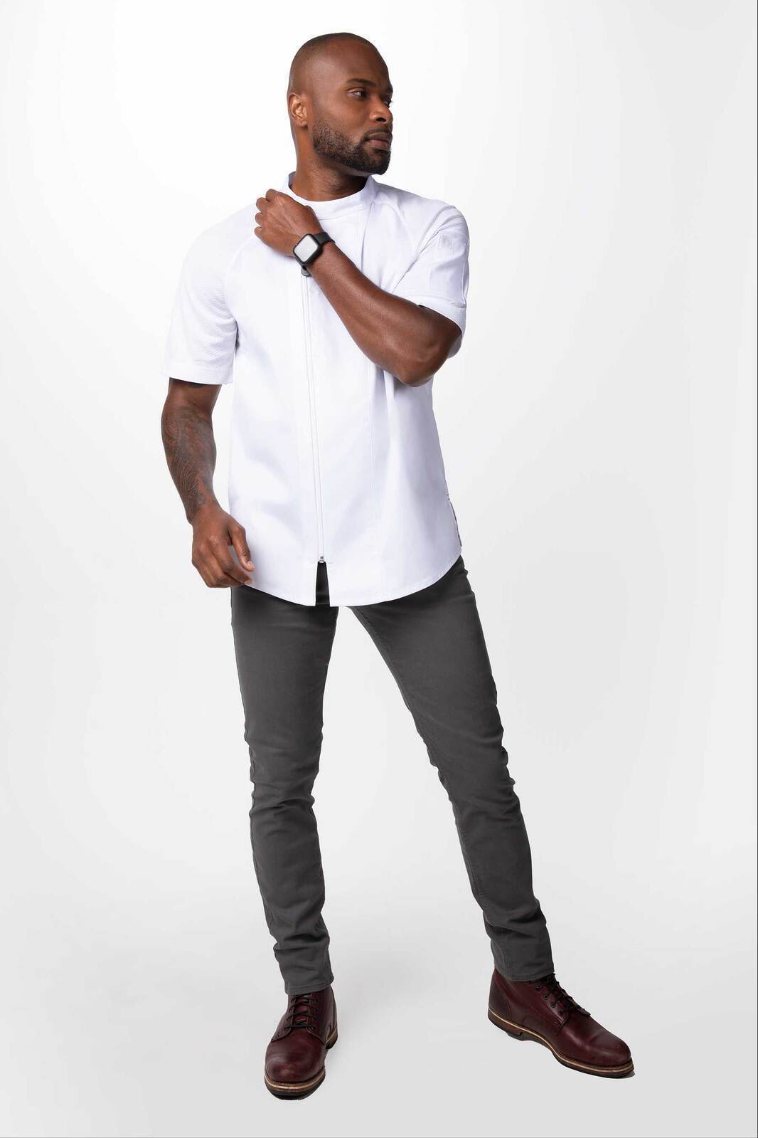 A man wearing a white chef jacket and grey pants, standing confidently with one hand on his shoulder.