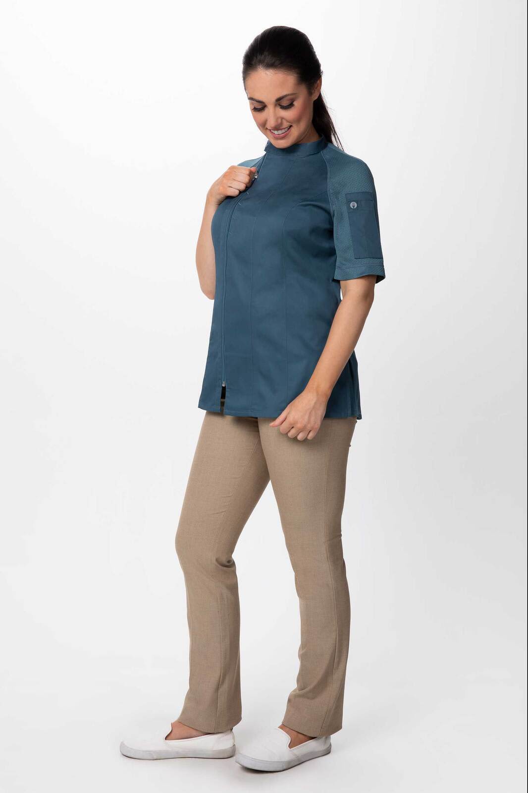 A woman is wearing a teal chef jacket coupled with light brown pants and white shoes. The jacket features a logo on the sleeve.