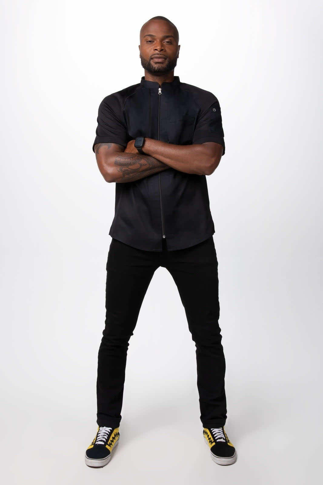 A black chef jacket worn by a man, paired with black pants and sneakers, standing confidently with arms crossed.