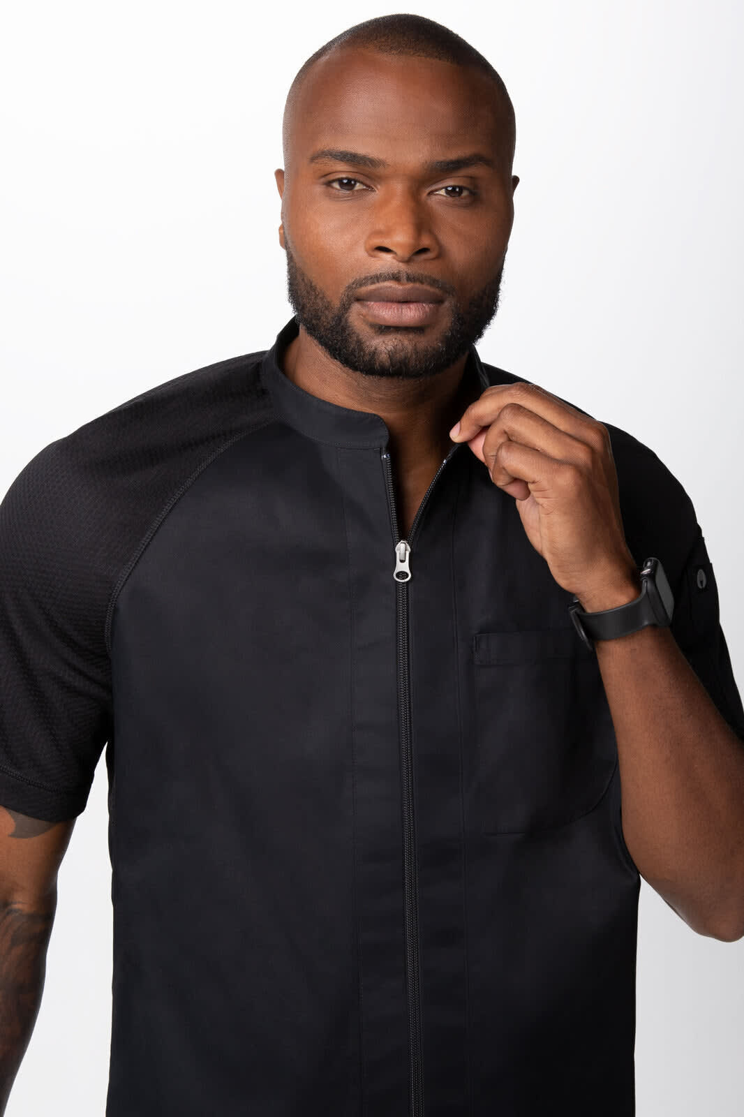 A black men's chef jacket is worn by a model, featuring a zip front and short sleeves. The model has a logo on the fabric.