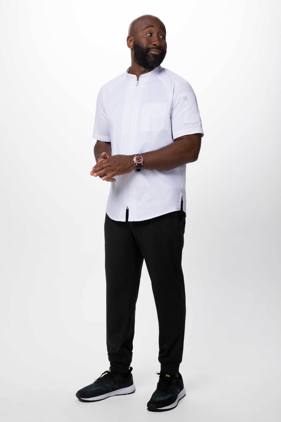 A man stands wearing a white chef jacket with a high collar and black pants, paired with athletic shoes.