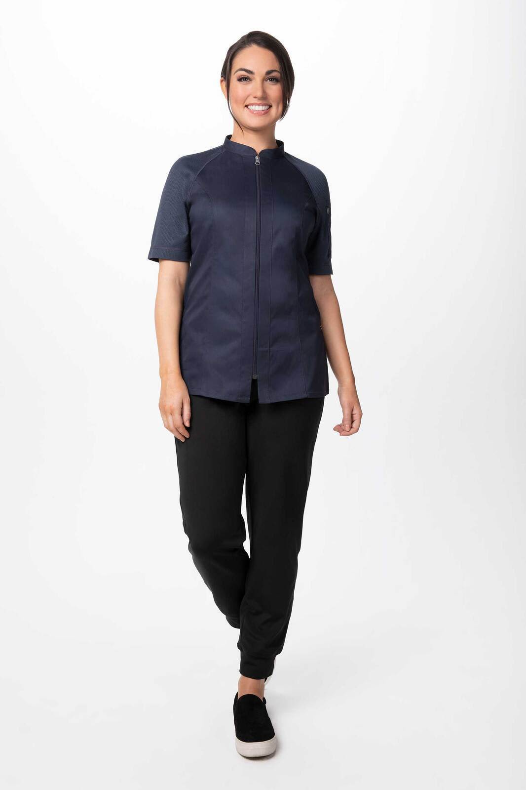 A woman wearing a navy blue chef jacket with short sleeves, paired with black pants and shoes. The jacket features a logo.