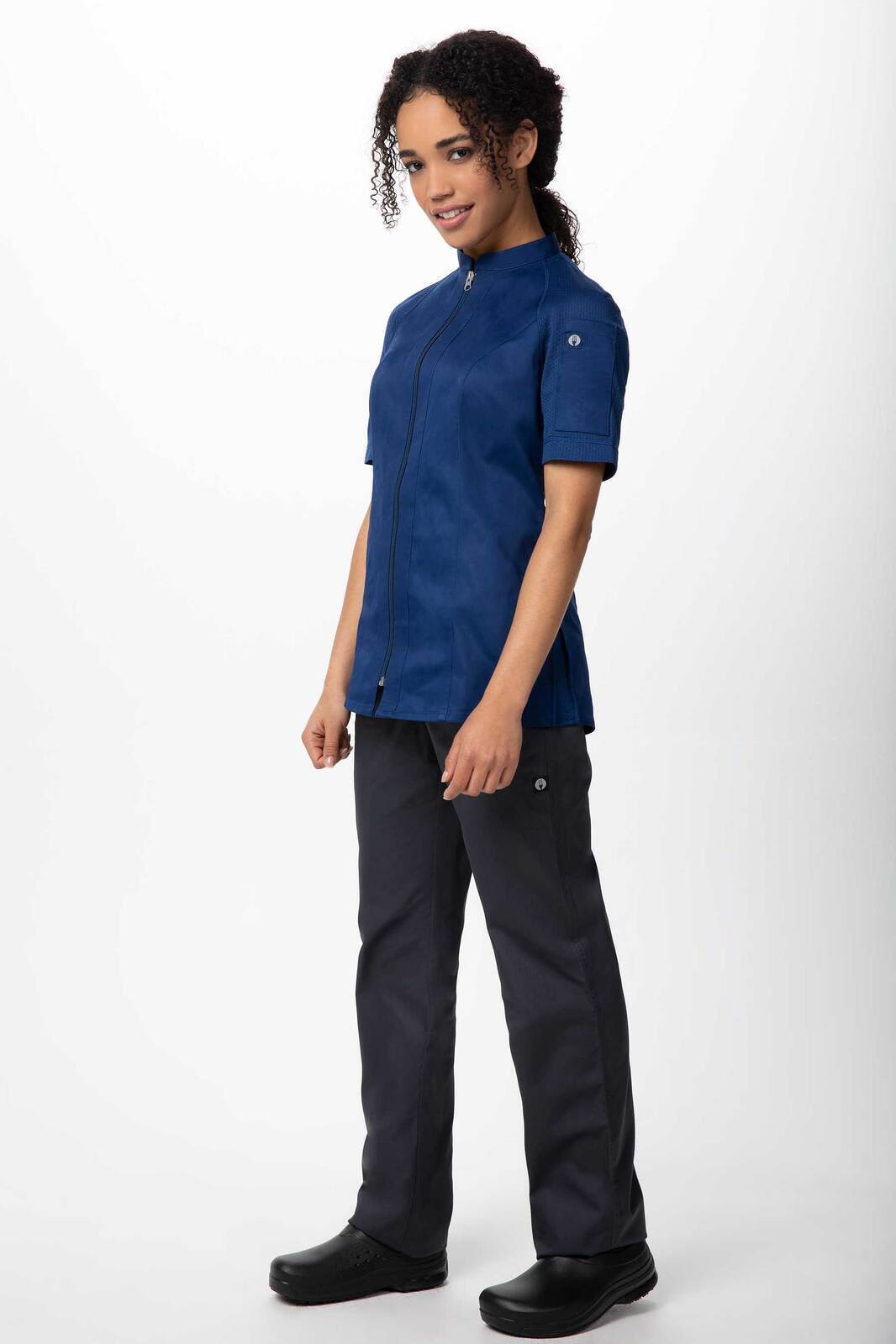 A woman stands in a navy blue chef jacket paired with dark grey pants and black shoes, featuring a logo.