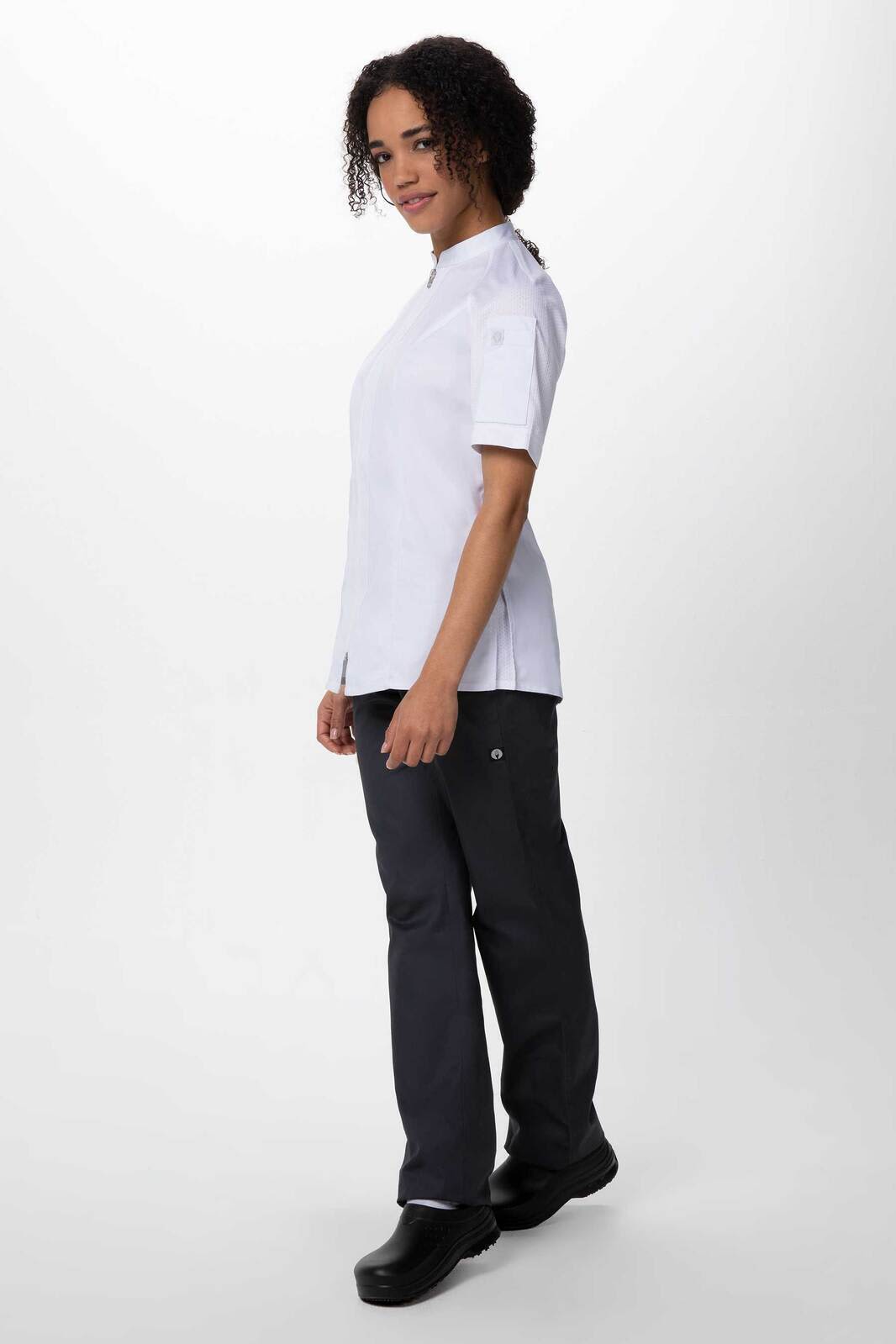 A woman stands wearing a white chef jacket and black chef pants, with a logo on the jacket.