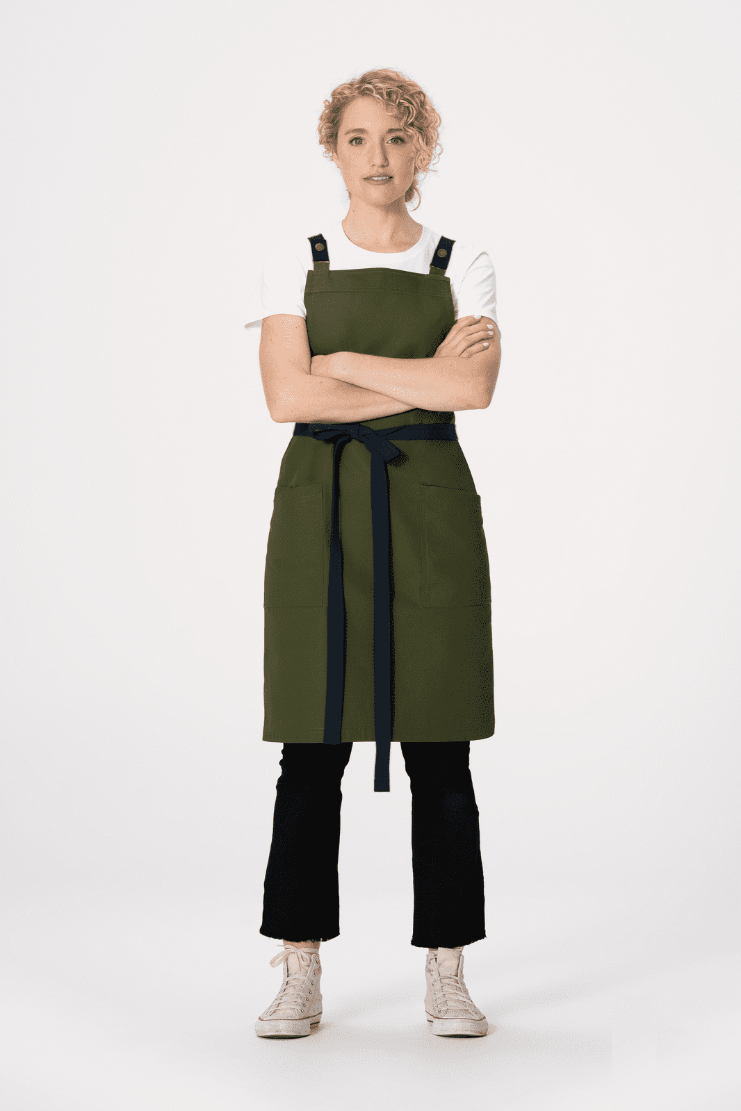 A green Lockharte cross back apron with black straps, worn by a model standing with arms crossed.