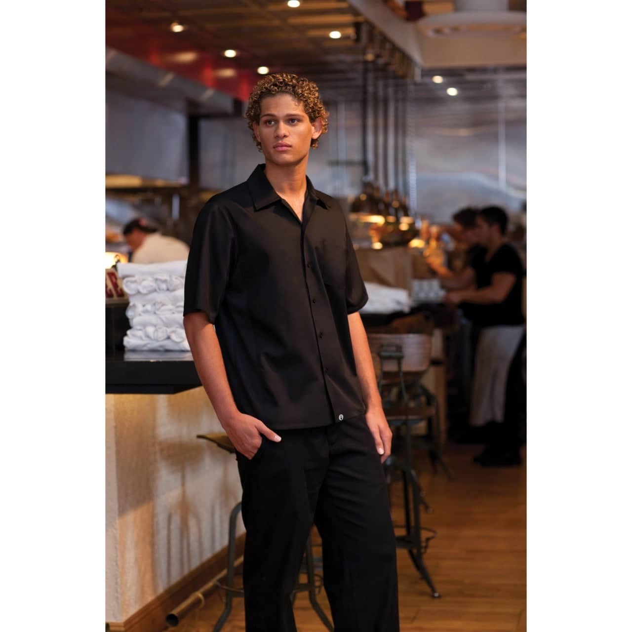 A black short-sleeve men's cook shirt displayed on a model in a restaurant setting with a logo present.
