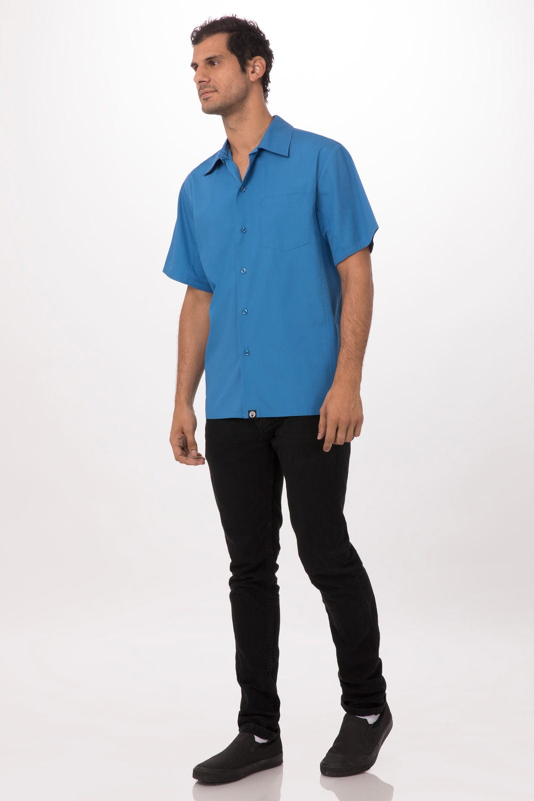 A man stands wearing a short-sleeve, blue shirt paired with black jeans and dark shoes. The shirt has a logo.