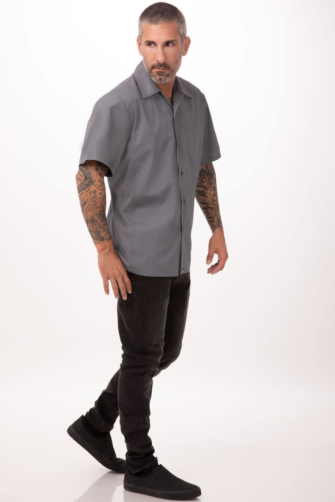 A grey short-sleeved men's shirt is displayed on a model walking, with tattoos on his arms and black trousers.