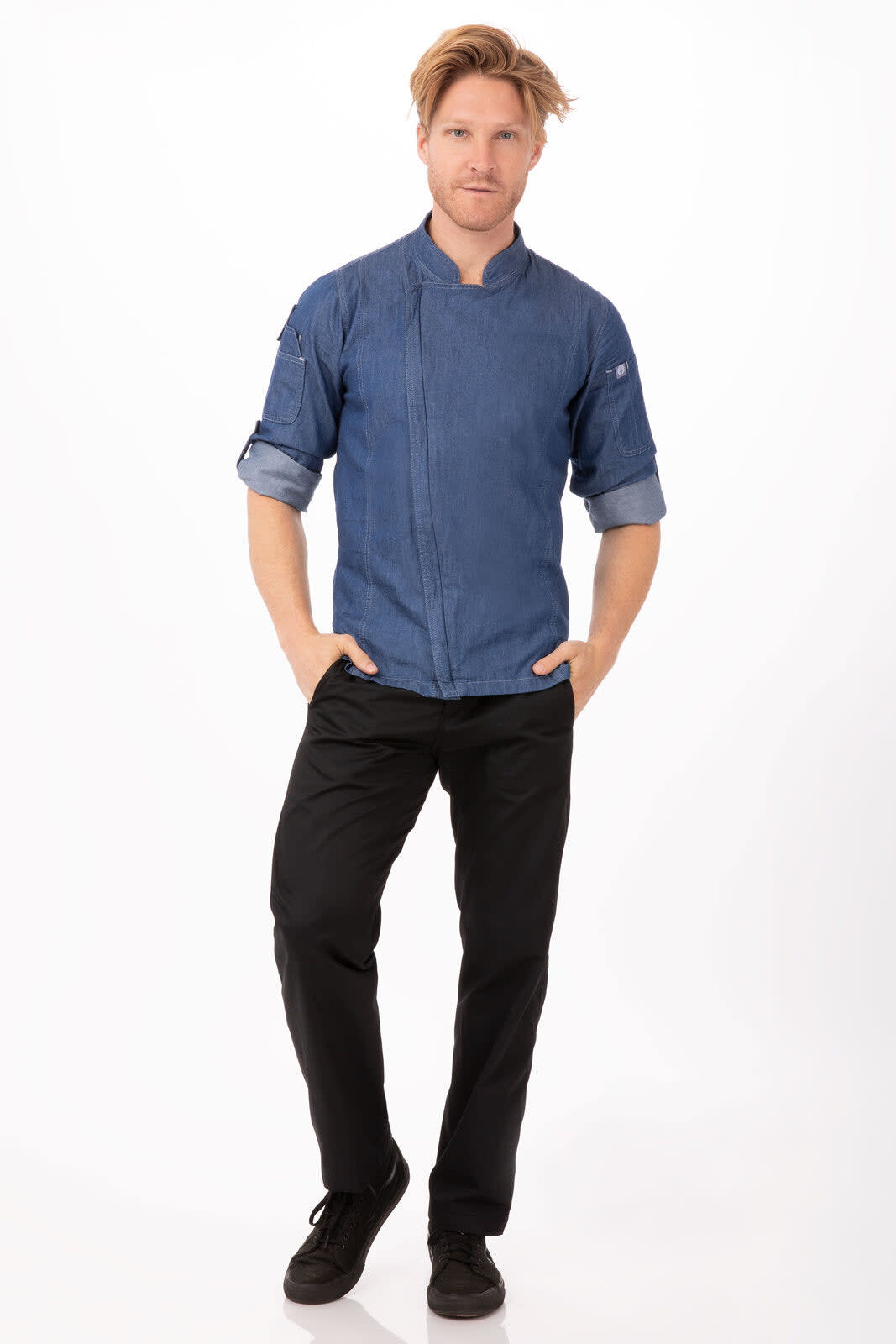 A man wearing a blue denim chef jacket and black pants, standing against a white background. The jacket features a logo.