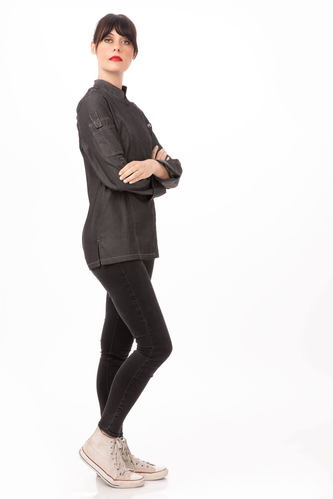A woman wearing a black denim chef jacket and black jeans, paired with light-coloured sneakers, standing confidently.