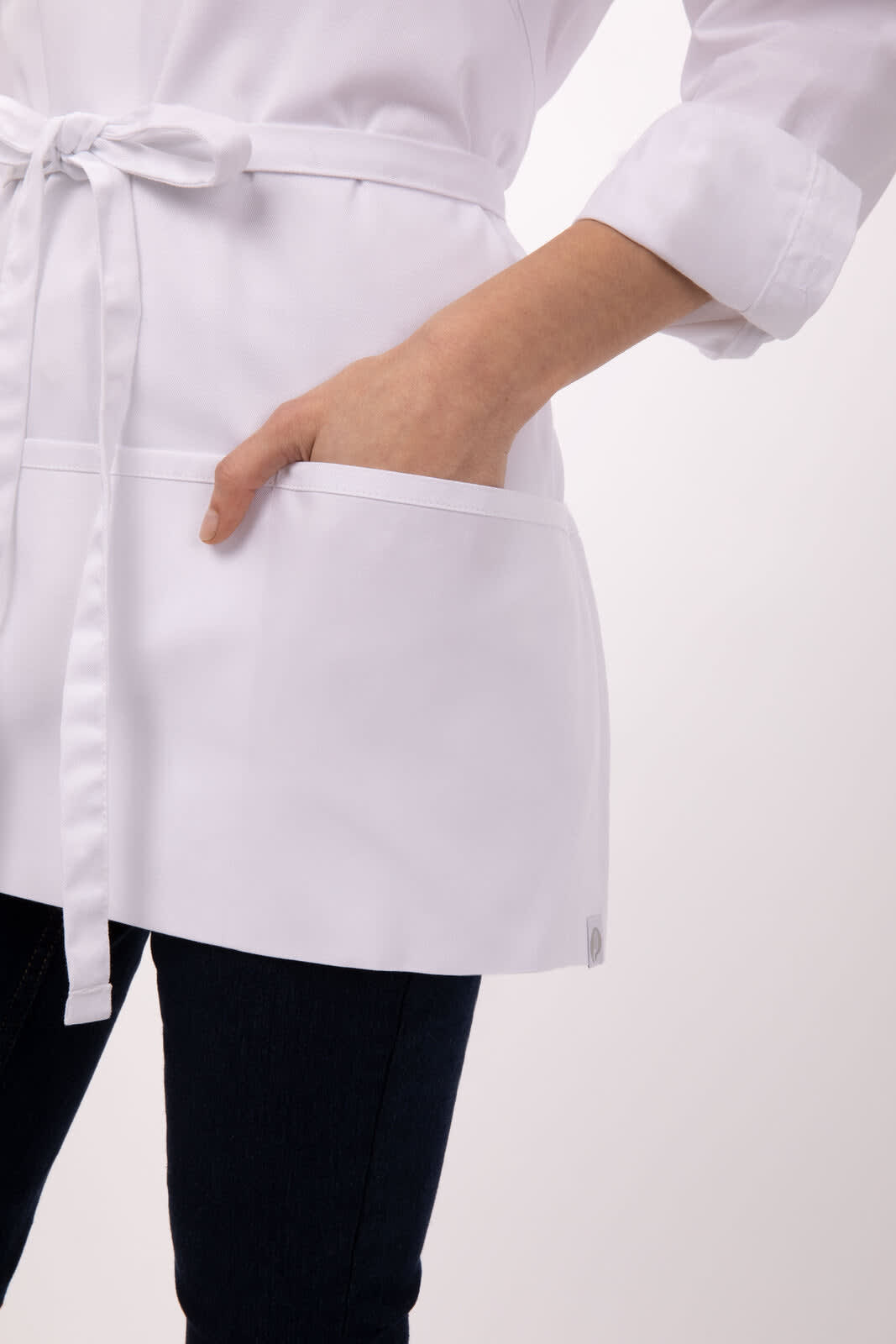 A white three-pocket apron with a tie waist and pockets visible on the front, worn over dark pants.