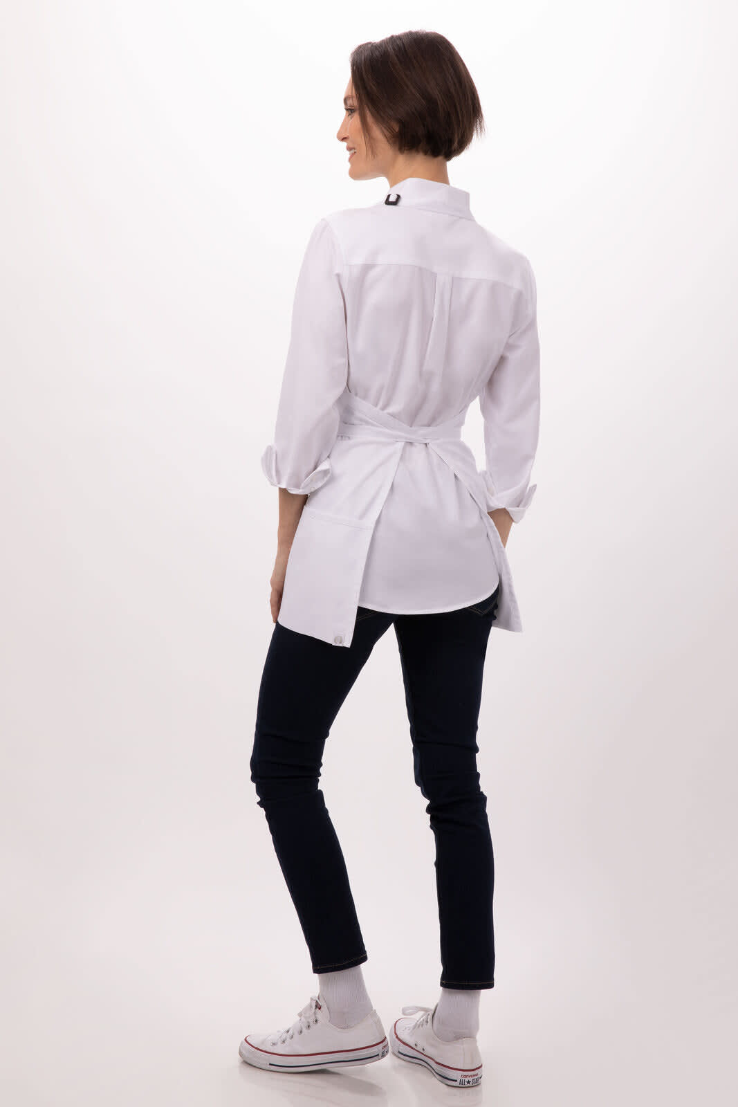 A three-pocket apron in white, worn backwards by a person, featuring a tie at the back. The model also wears dark pants and white shoes.