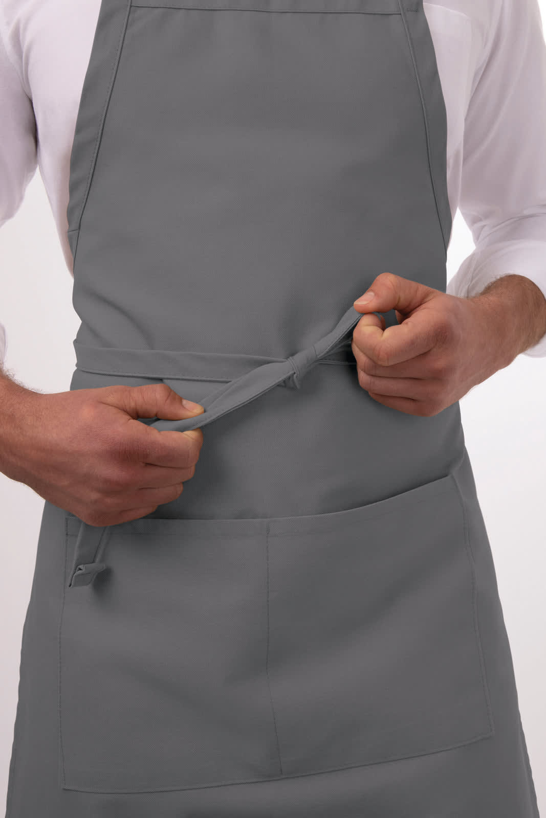 A grey bib apron featuring a front pocket, held by a person adjusting the ties. The product has a minimalist design.