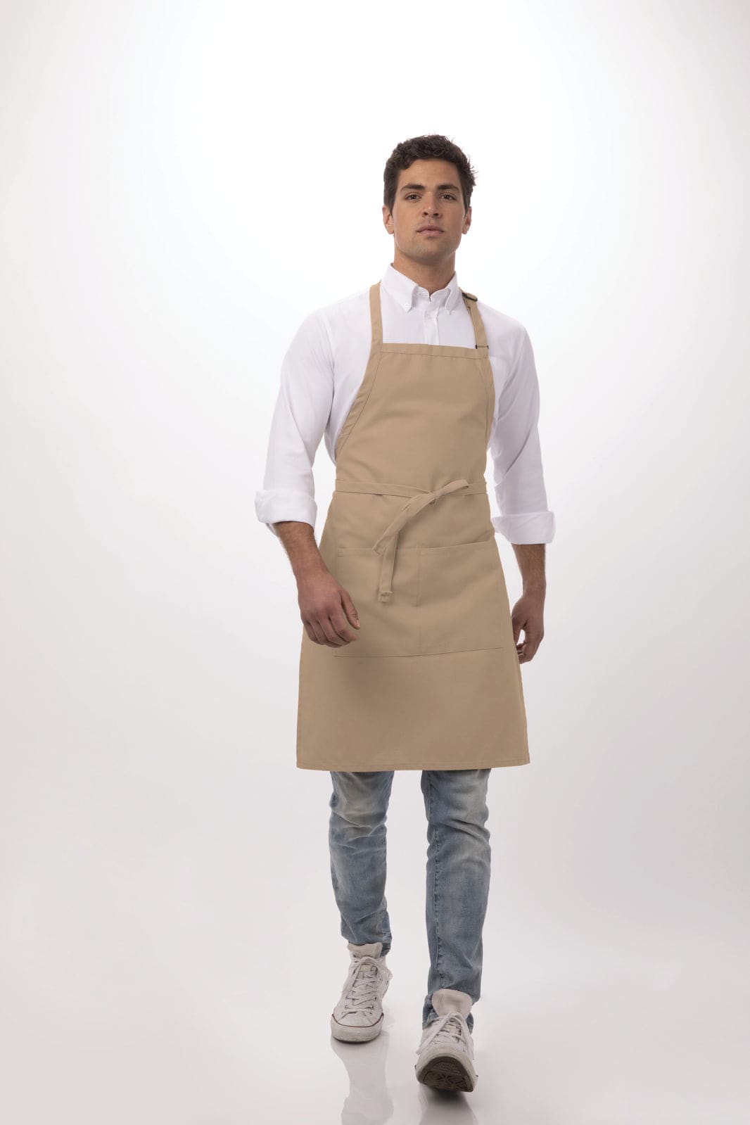 A beige bib apron is worn by a model over a white shirt and light jeans. The apron features a front pocket and has a logo.