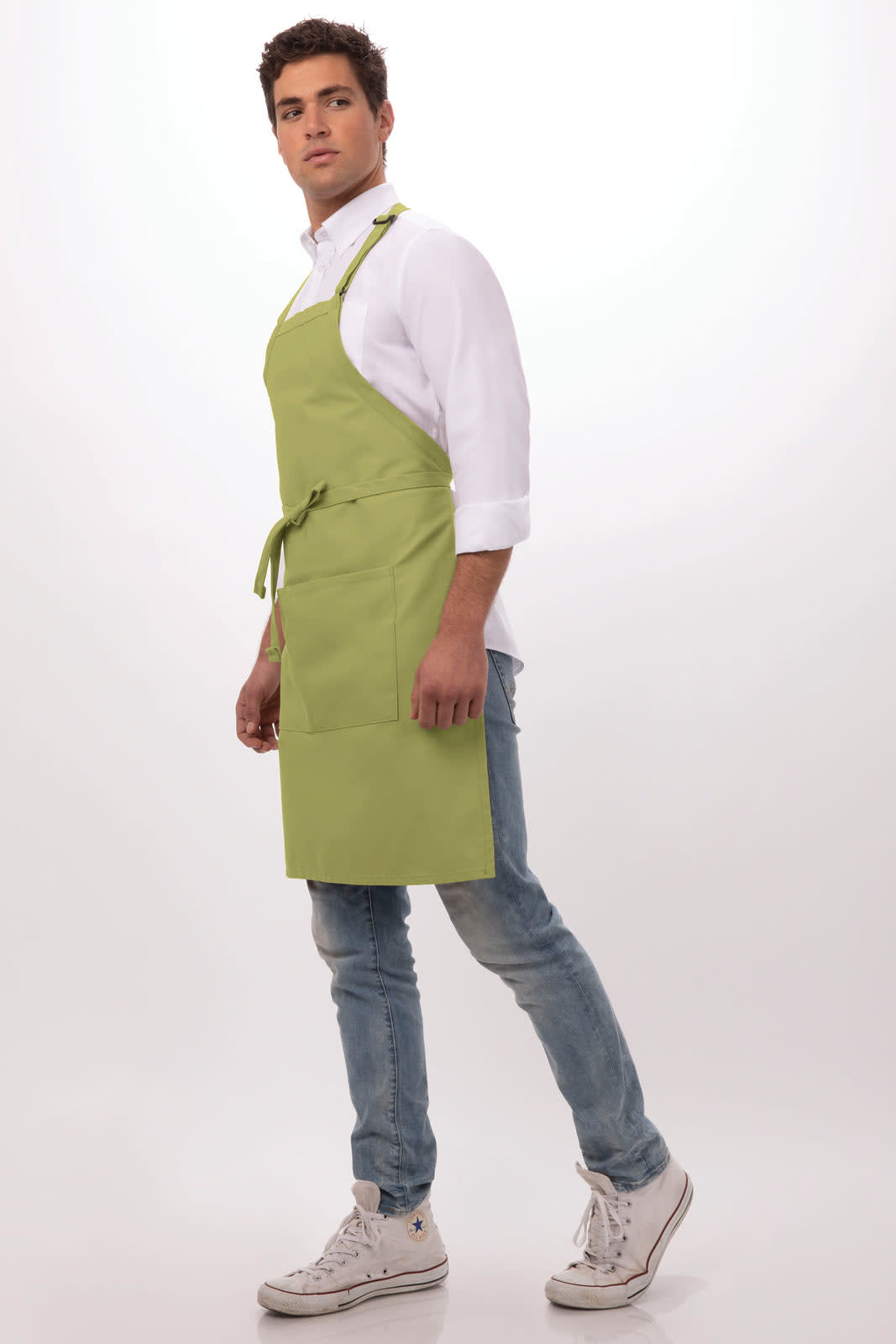 A man wearing a light green bib apron over a white shirt, styled with blue jeans and sneakers, featuring a logo.