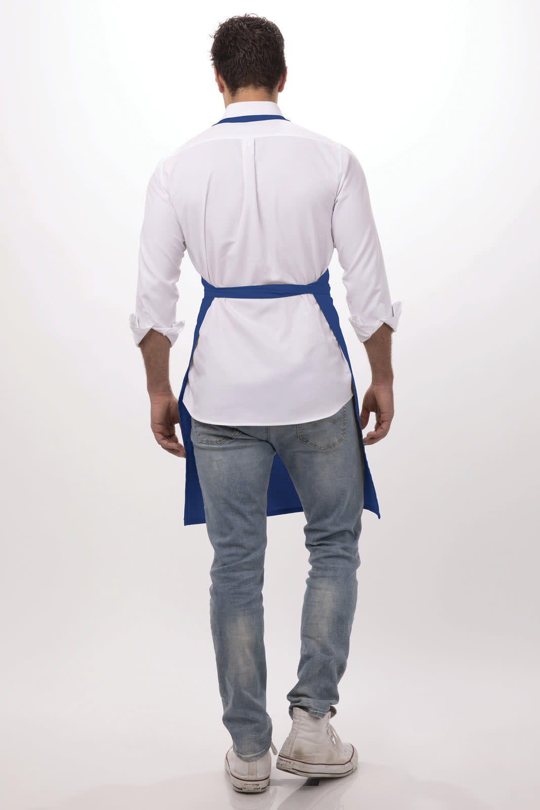 A man wearing a white shirt and blue bib apron, viewed from the back, paired with light denim jeans and white sneakers.