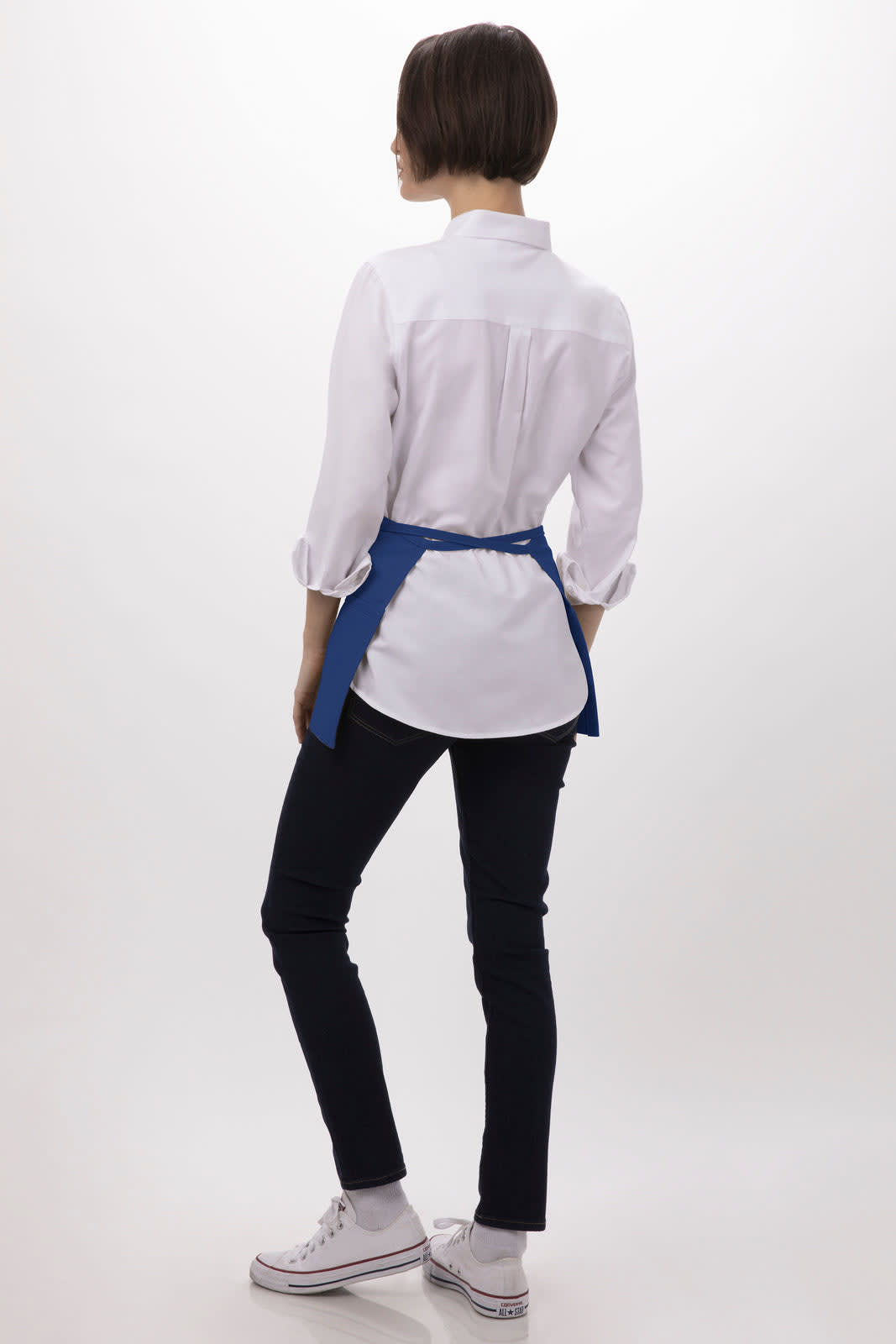 A waist apron in blue, worn over a white shirt, paired with dark trousers and white sneakers. The model is facing away.