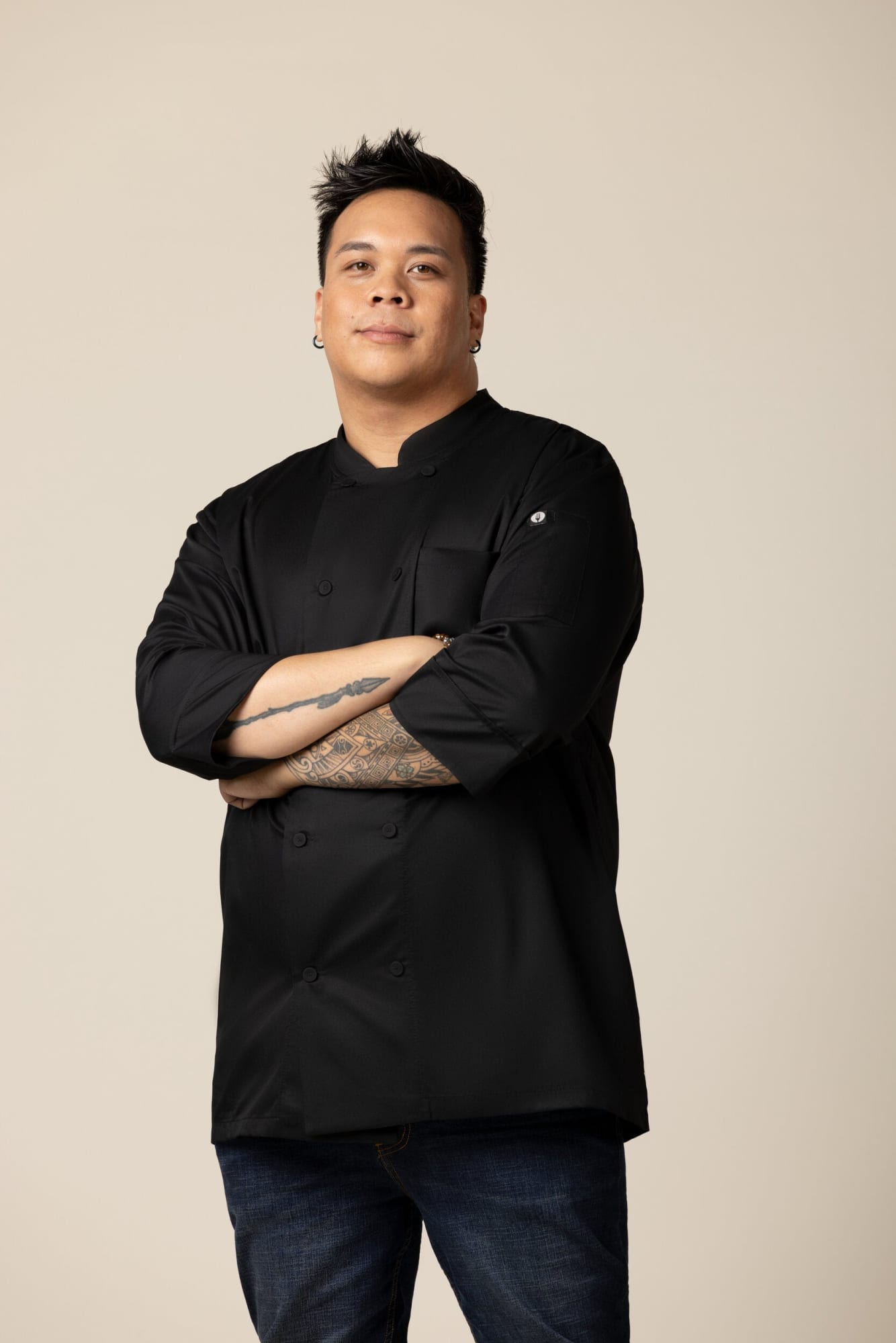 A man wearing a black chef jacket stands confidently with arms crossed, showcasing the attire against a neutral background.