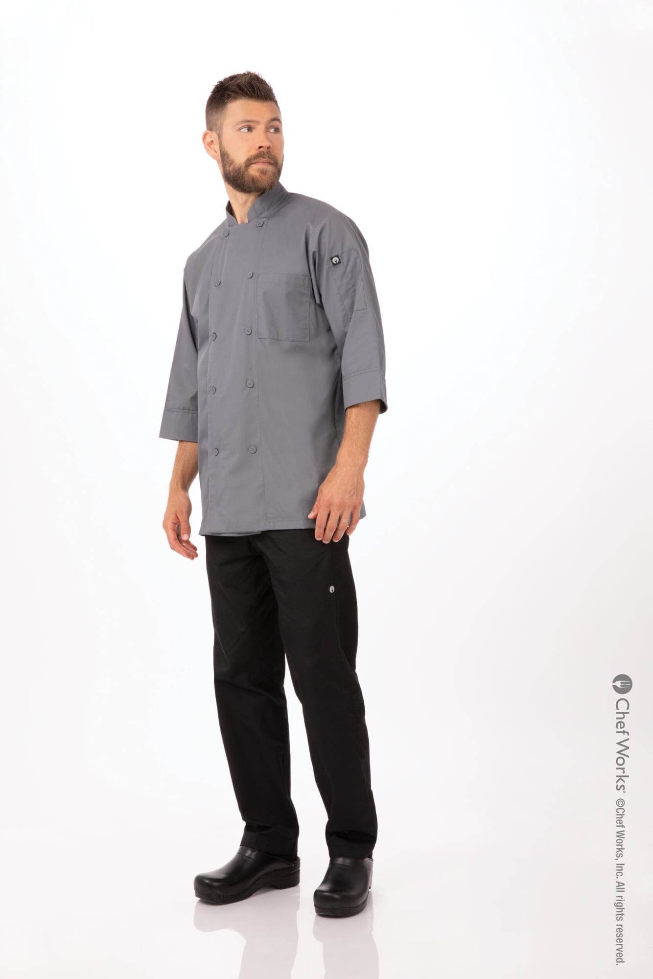 A man wearing a grey chef jacket and black pants, standing against a plain background. The jacket features a logo.