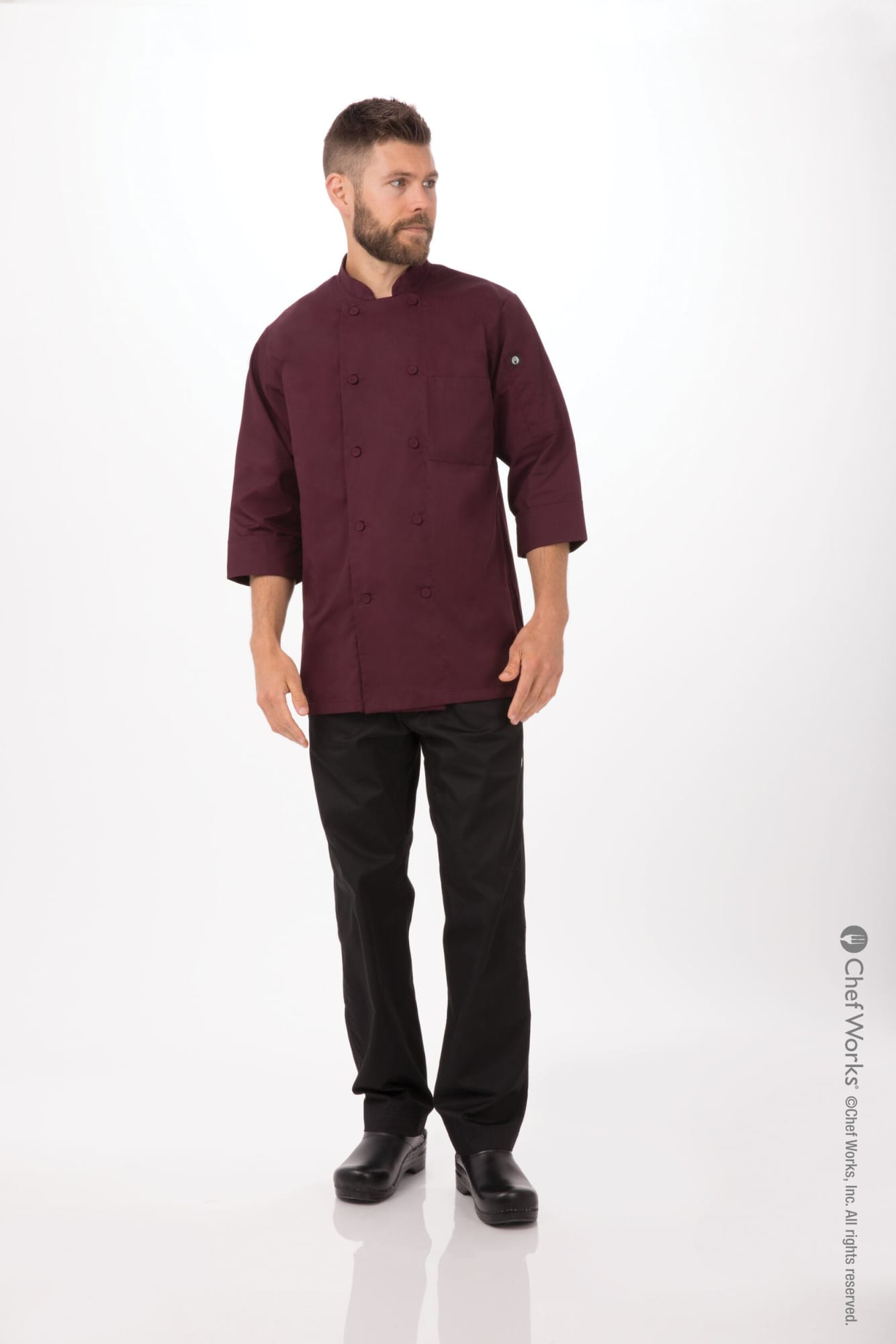 A man wearing a burgundy men's chef jacket and black pants, standing against a light background. The jacket features a logo.