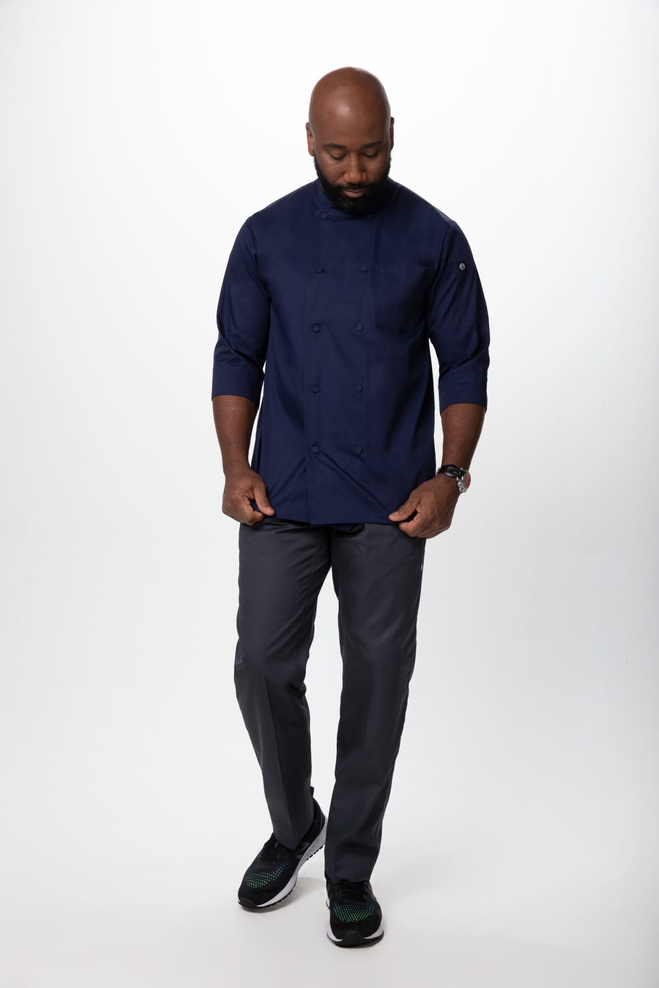 A men's chef jacket in navy blue, paired with grey trousers, worn by a man standing and walking. The jacket has a logo.