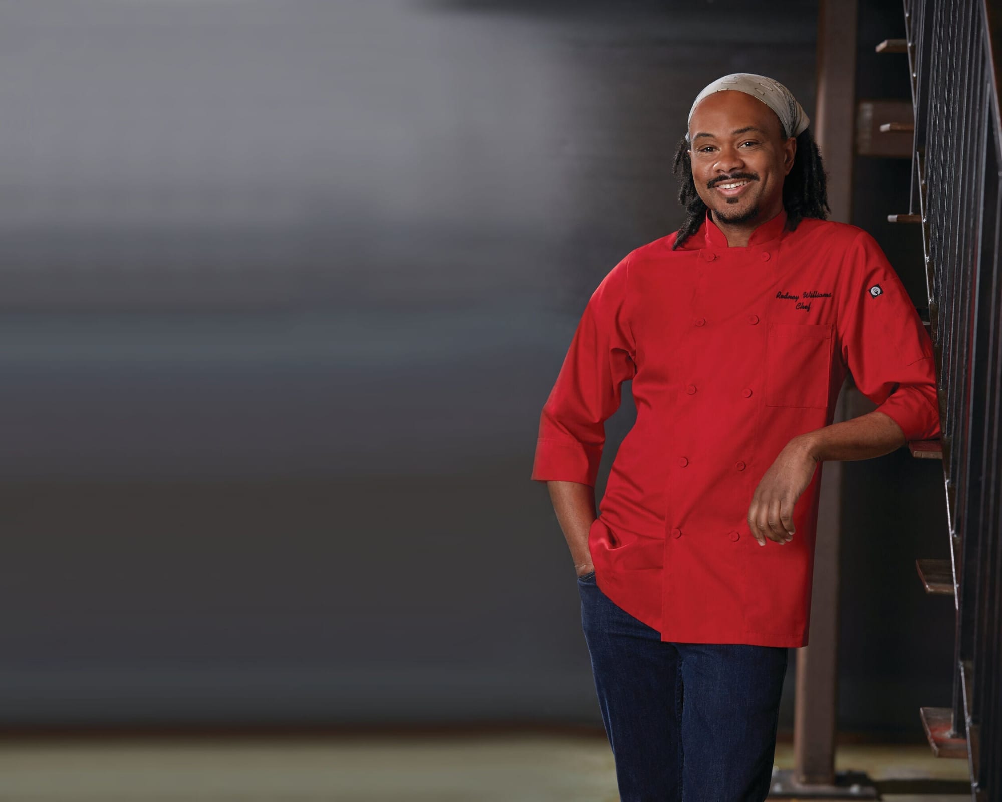 A male model wearing a red chef jacket with a light-coloured headscarf, leaning against a railing.