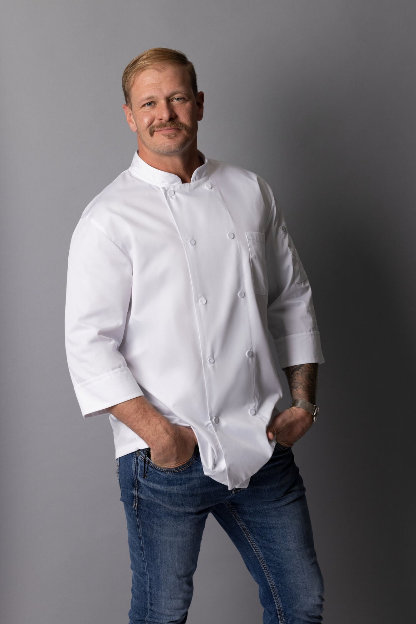 A man wearing a white chef jacket with double-breasted buttons, paired with blue jeans, stands casually.