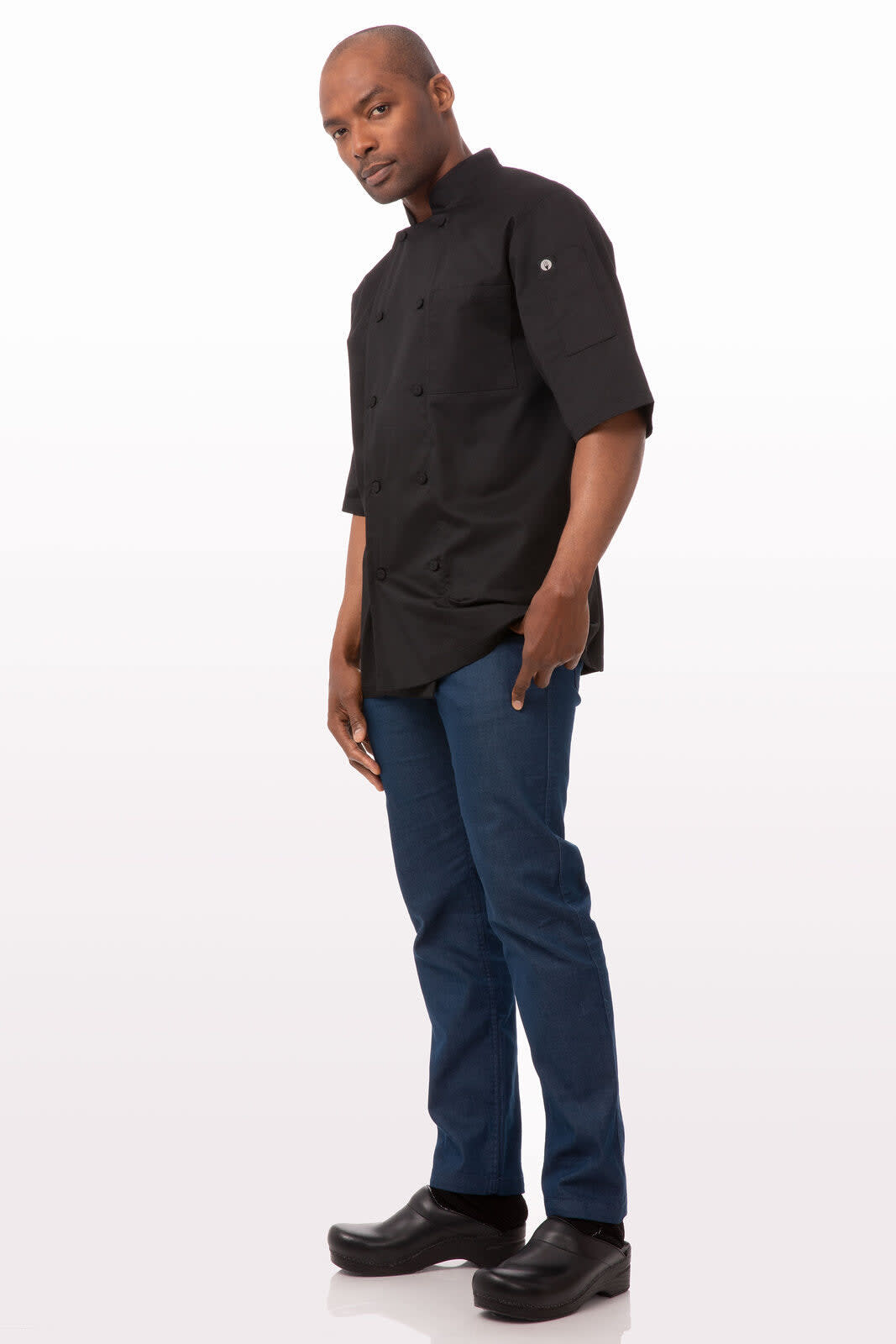 A black chef jacket is worn by a man paired with dark blue jeans and black footwear. The jacket has a logo.